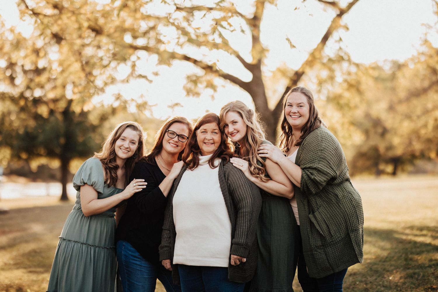 Five women spanning generations smiling together under golden trees in a park setting.