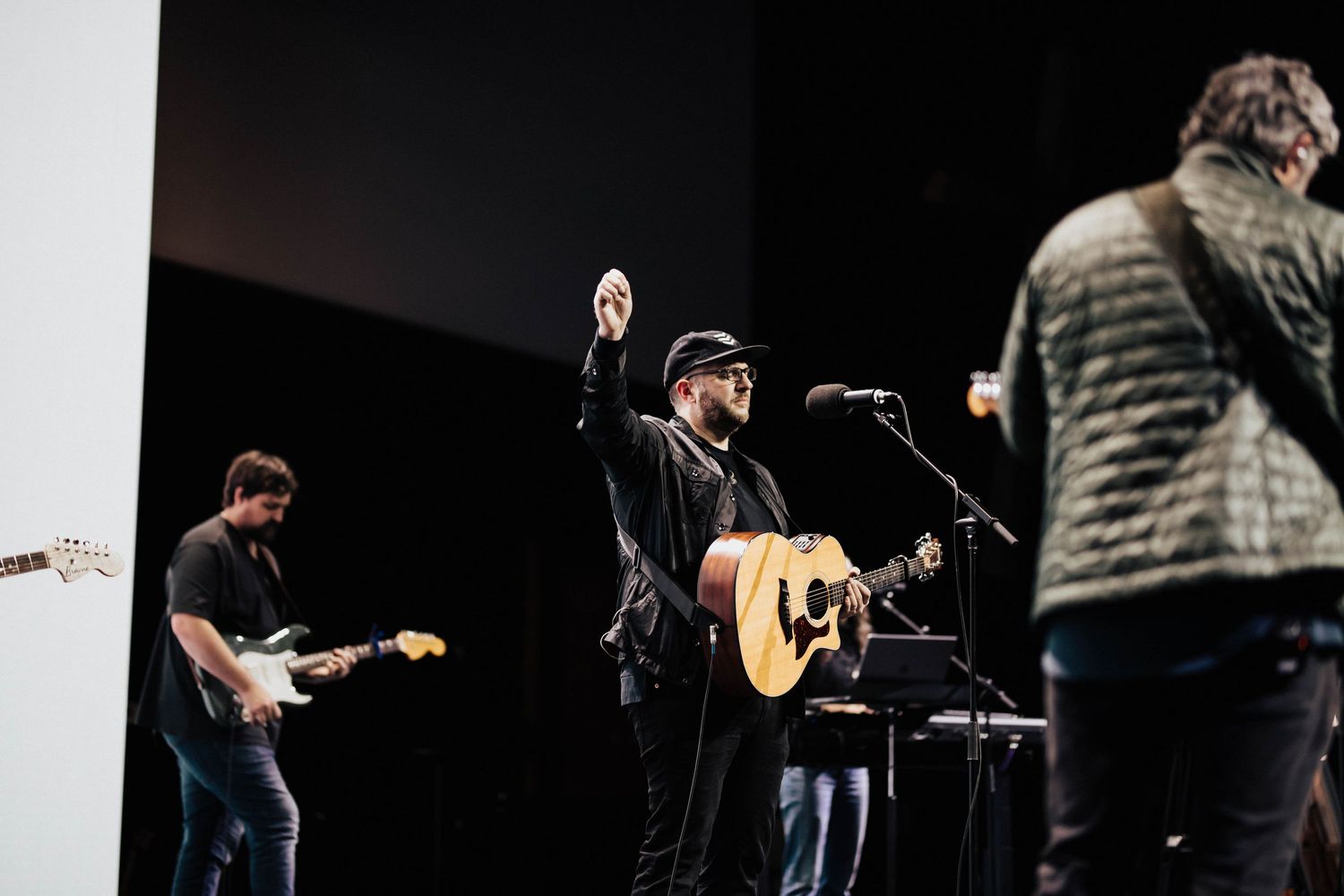 Band performing on stage with guitarist, vocalist raising hand holding mic, and acoustic guitar player under dramatic lighting.