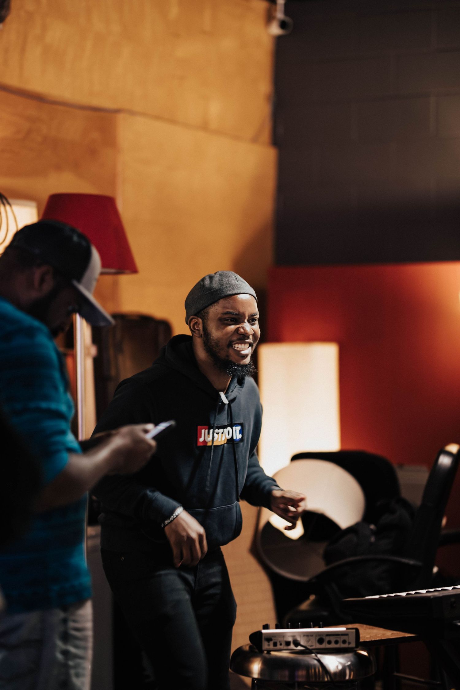 Two men laugh and converse in a warmly lit recording studio with red accents and musical equipment nearby.