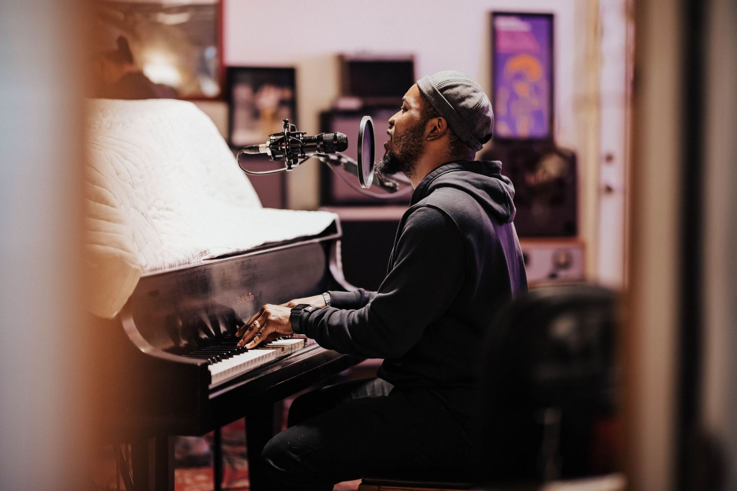 Musician singing into a microphone while playing an upright piano in a cozy studio.