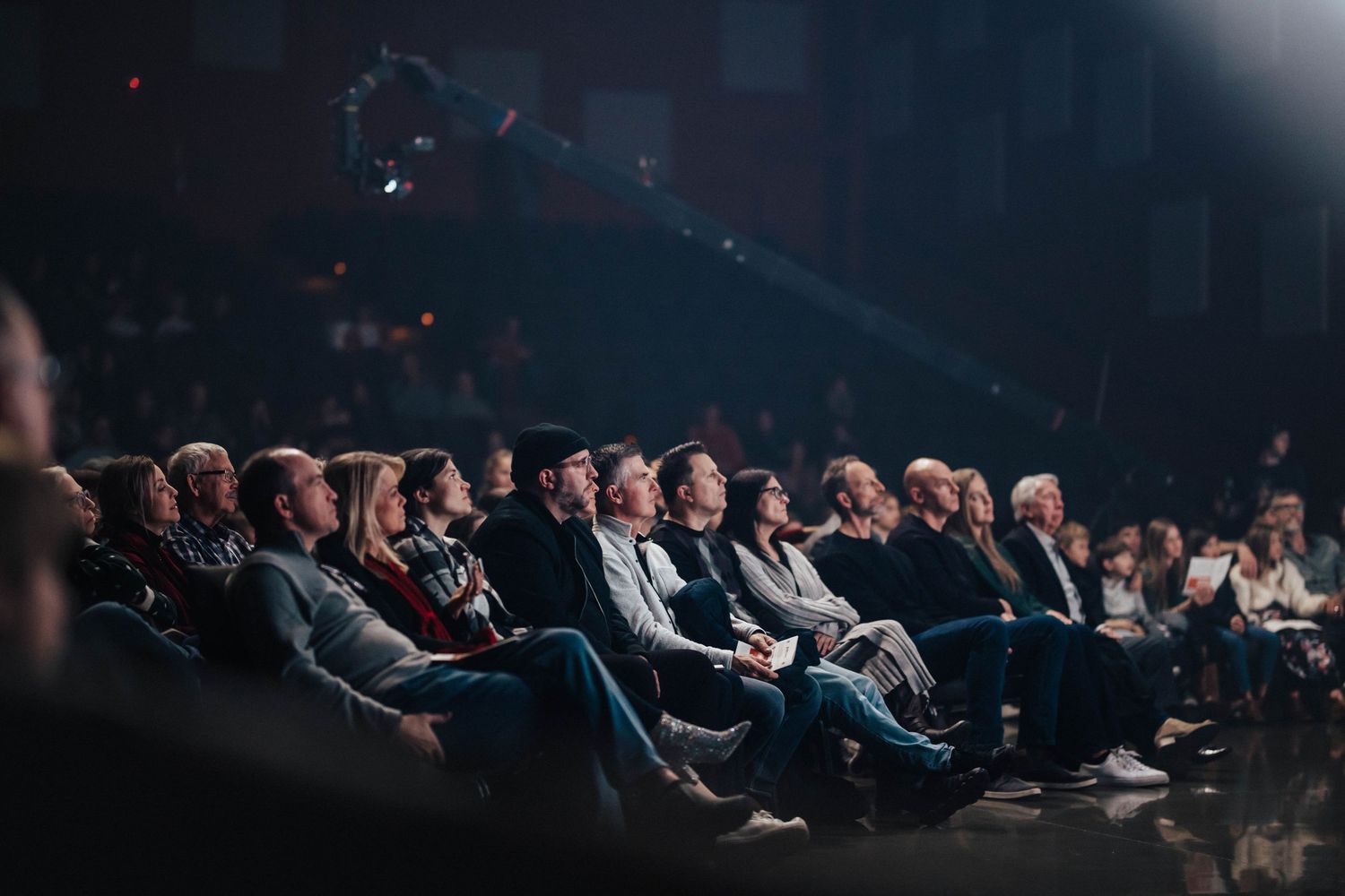 Audience seated in darkened theater venue, illuminated by stage lighting during a live event.