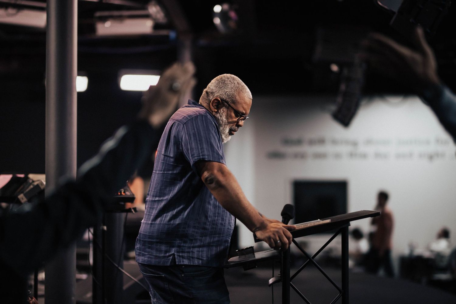 An elderly man with white hair and glasses leans on a railing inside a dimly lit church venue.