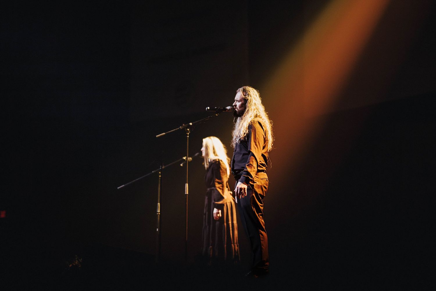 Two performers with long hair stand together at a microphone on a dimly lit stage with warm amber lighting.