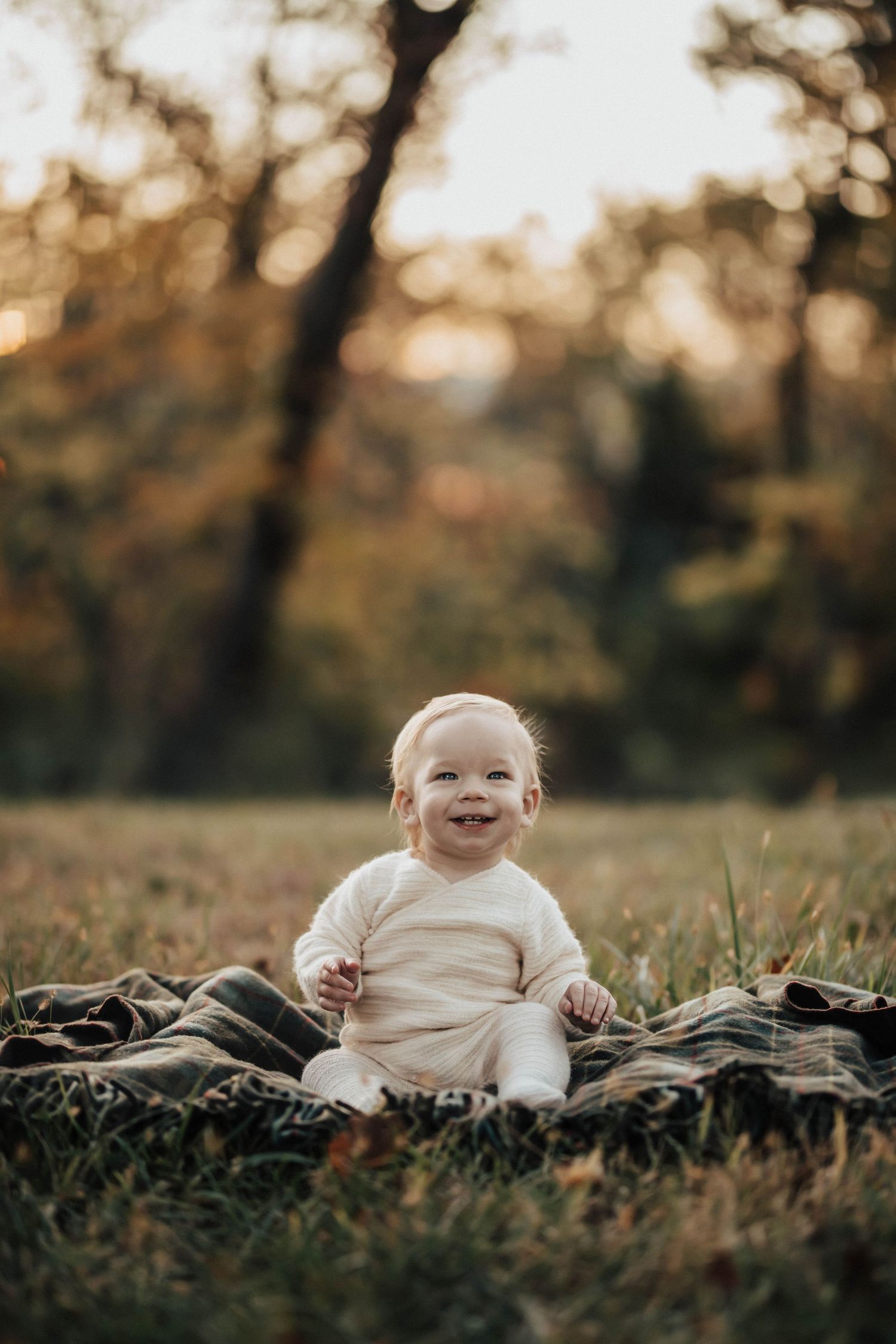 Smiling baby sitting on a plaid blanket in a sunlit autumn park, wearing a white outfit.