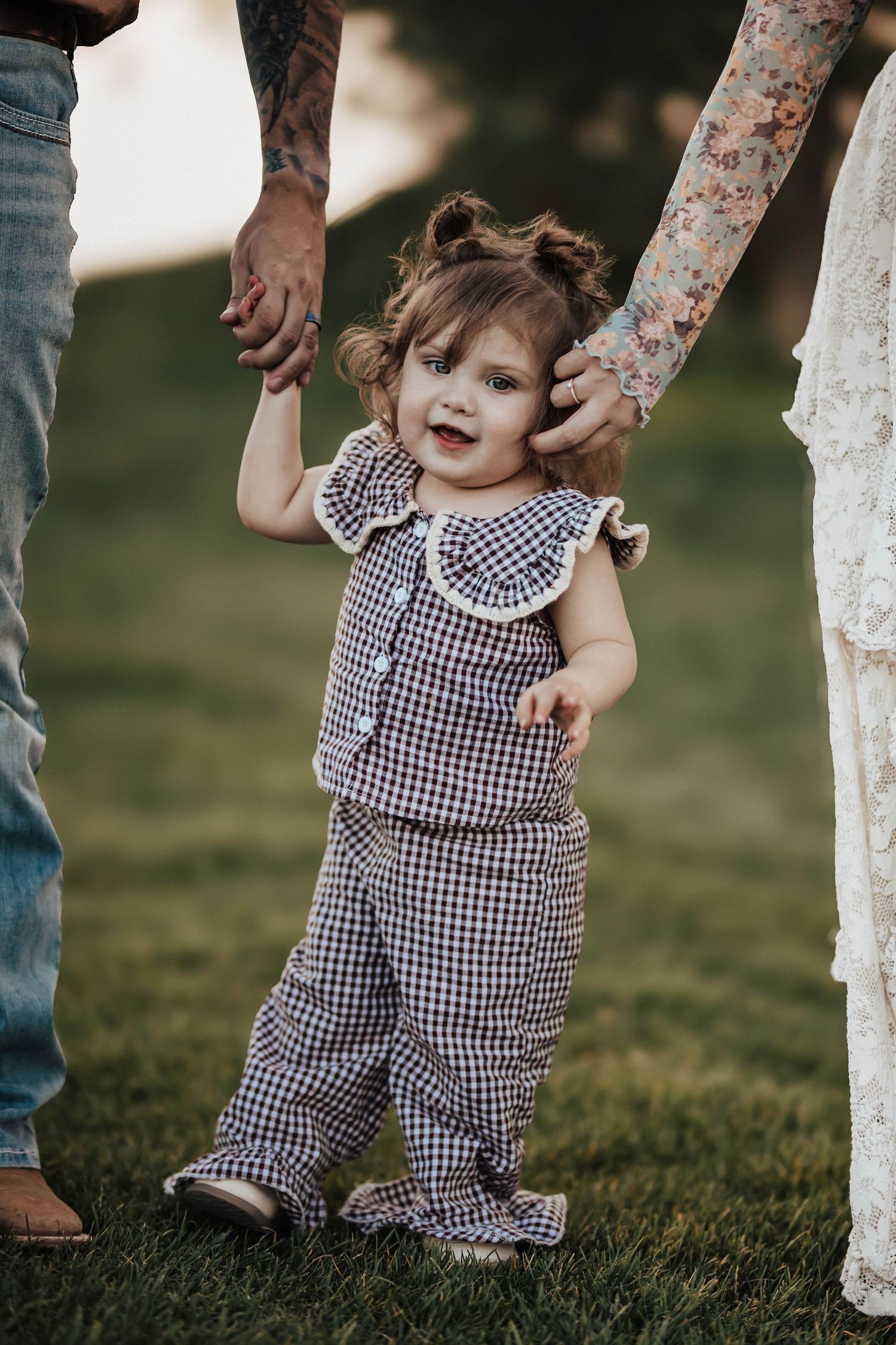 Toddler girl in gingham romper smiling while holding hands with parents on green grass.