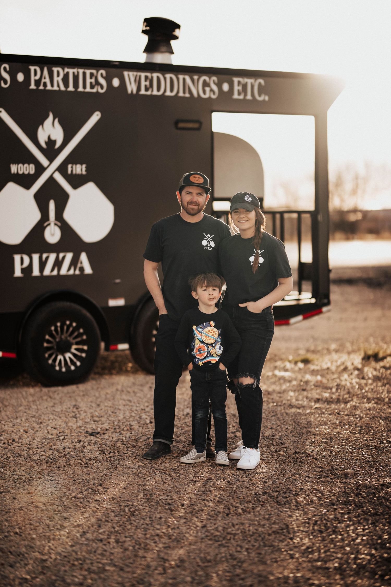 Family of three poses in front of a black Wood Fire Pizza catering trailer at sunset.