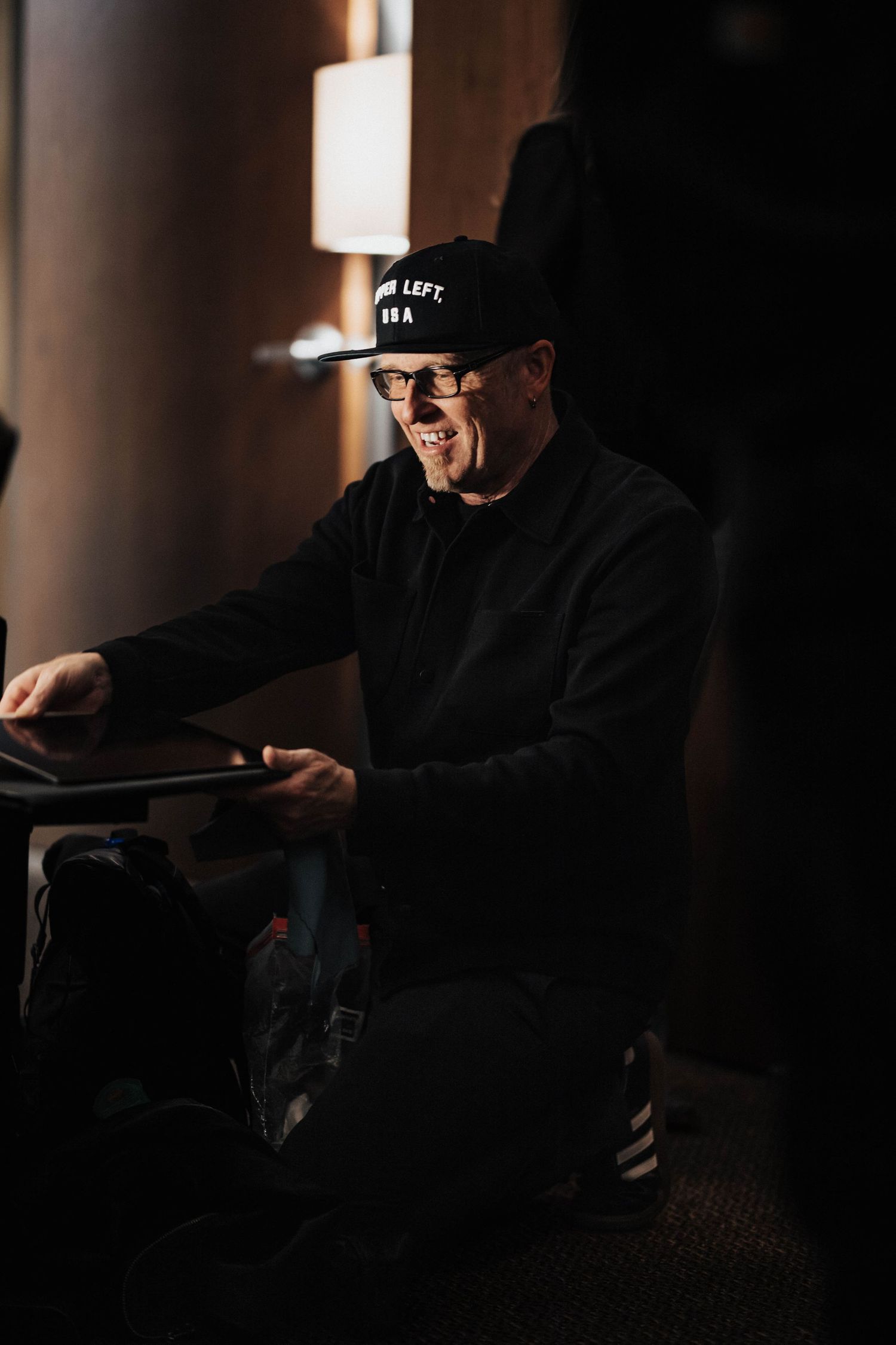 Smiling man in black cap and glasses seated at table in warmly lit backstage setting at music event.