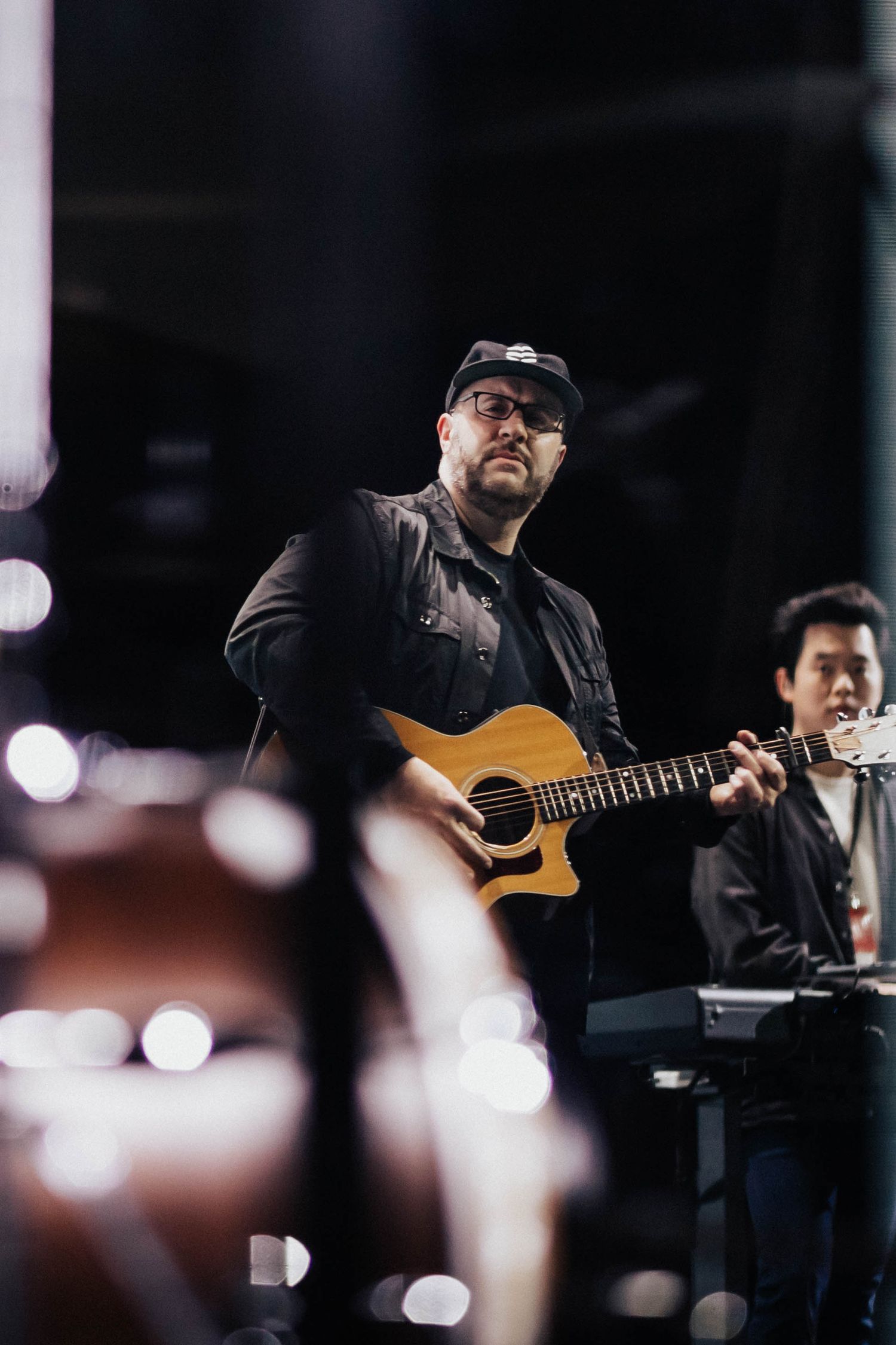 Man in black cap playing acoustic guitar on stage with keyboard player in background at live concert.
