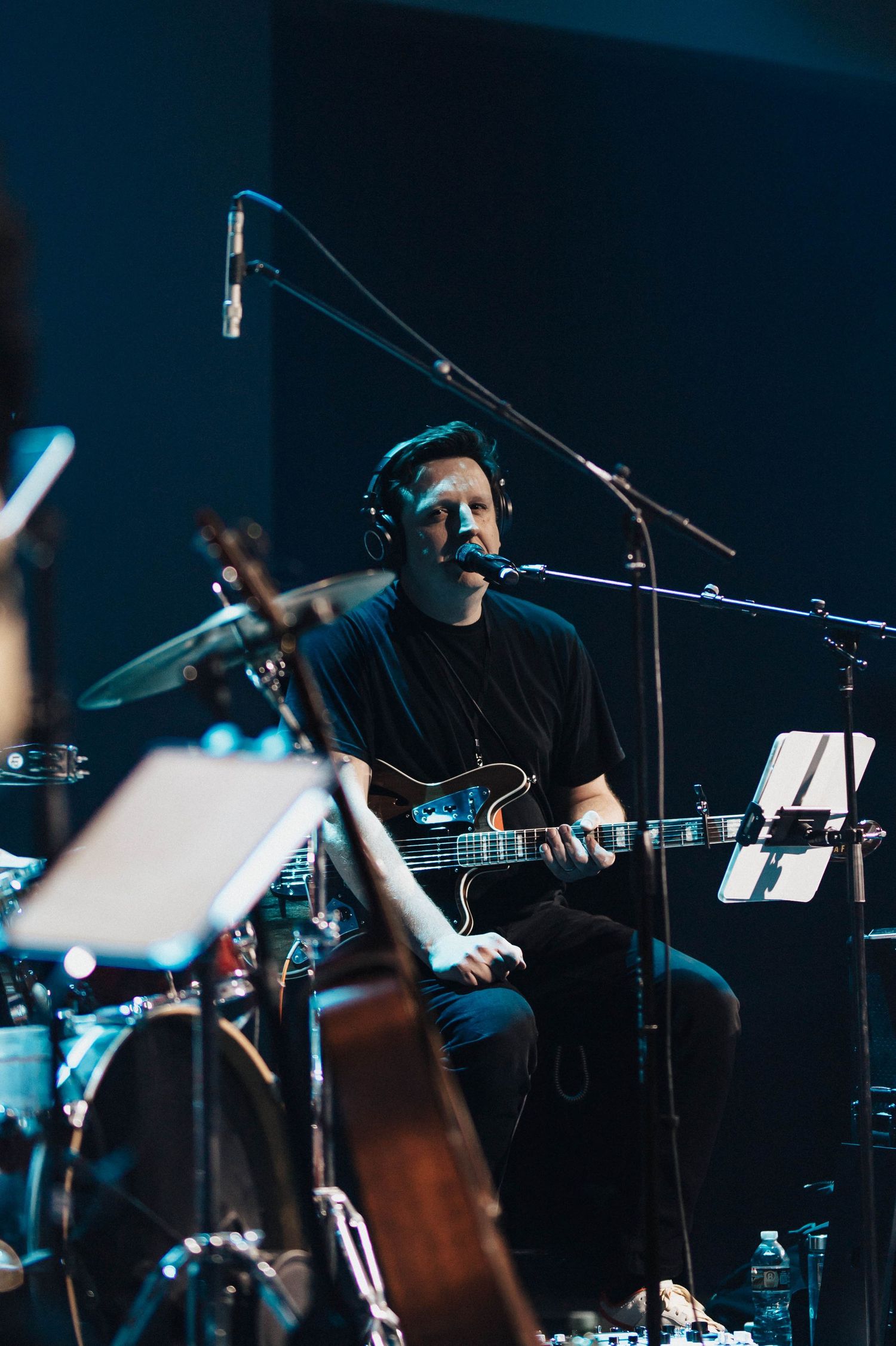 Male musician playing electric guitar and singing into a microphone in a dark recording studio.