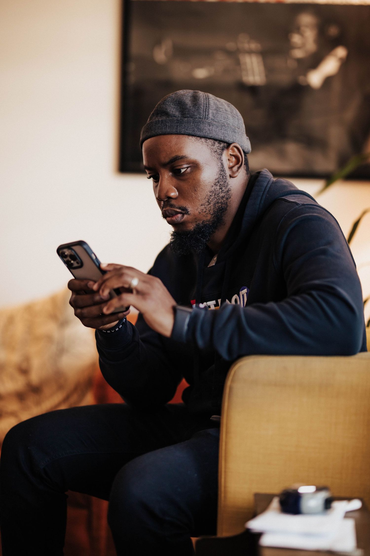 Young Black man in gray beanie and navy hoodie sitting in chair, focused on smartphone screen indoors.