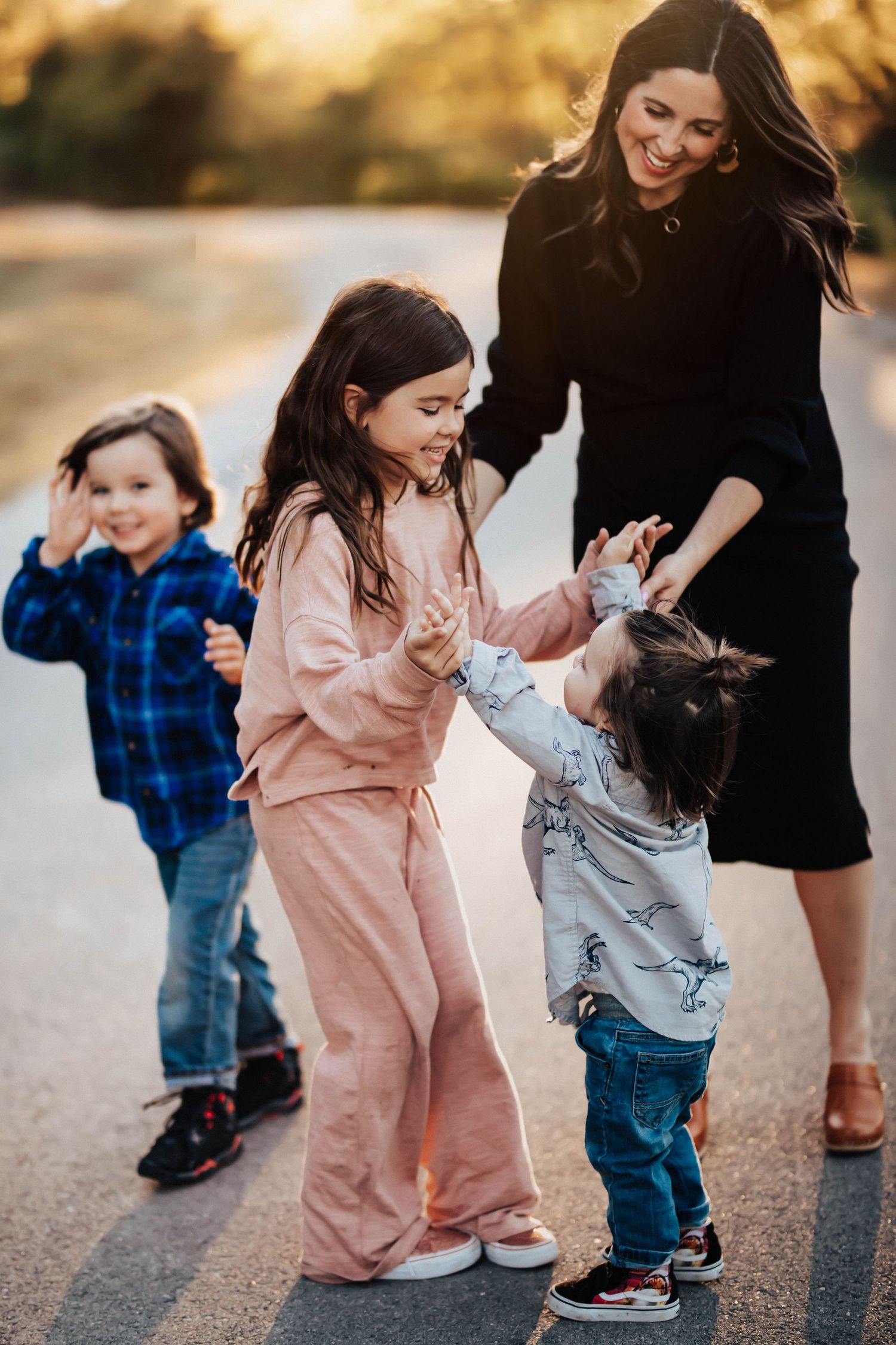 A smiling mother in black dress watches her three young children dance and play on a road.