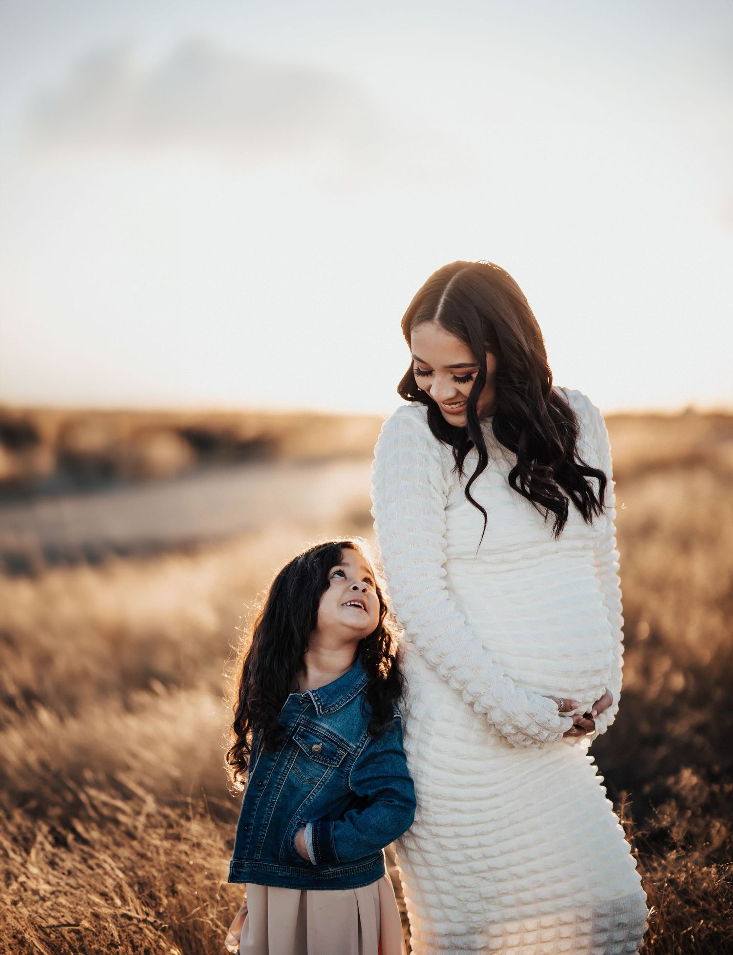 Pregnant woman in white dress with young girl in field at golden hour.