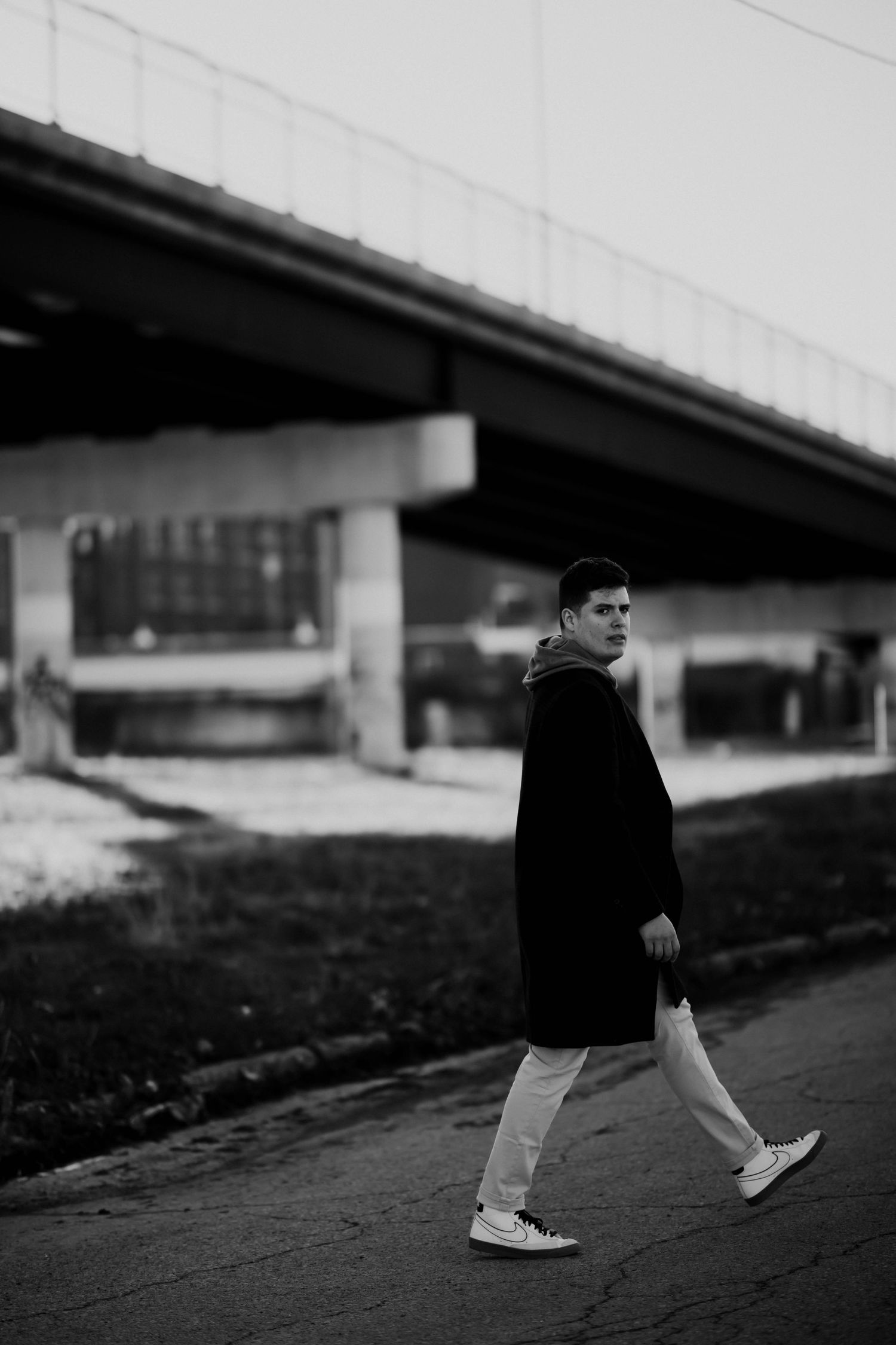 Young man in black coat and white pants walks near bridge underpass in black and white photo.