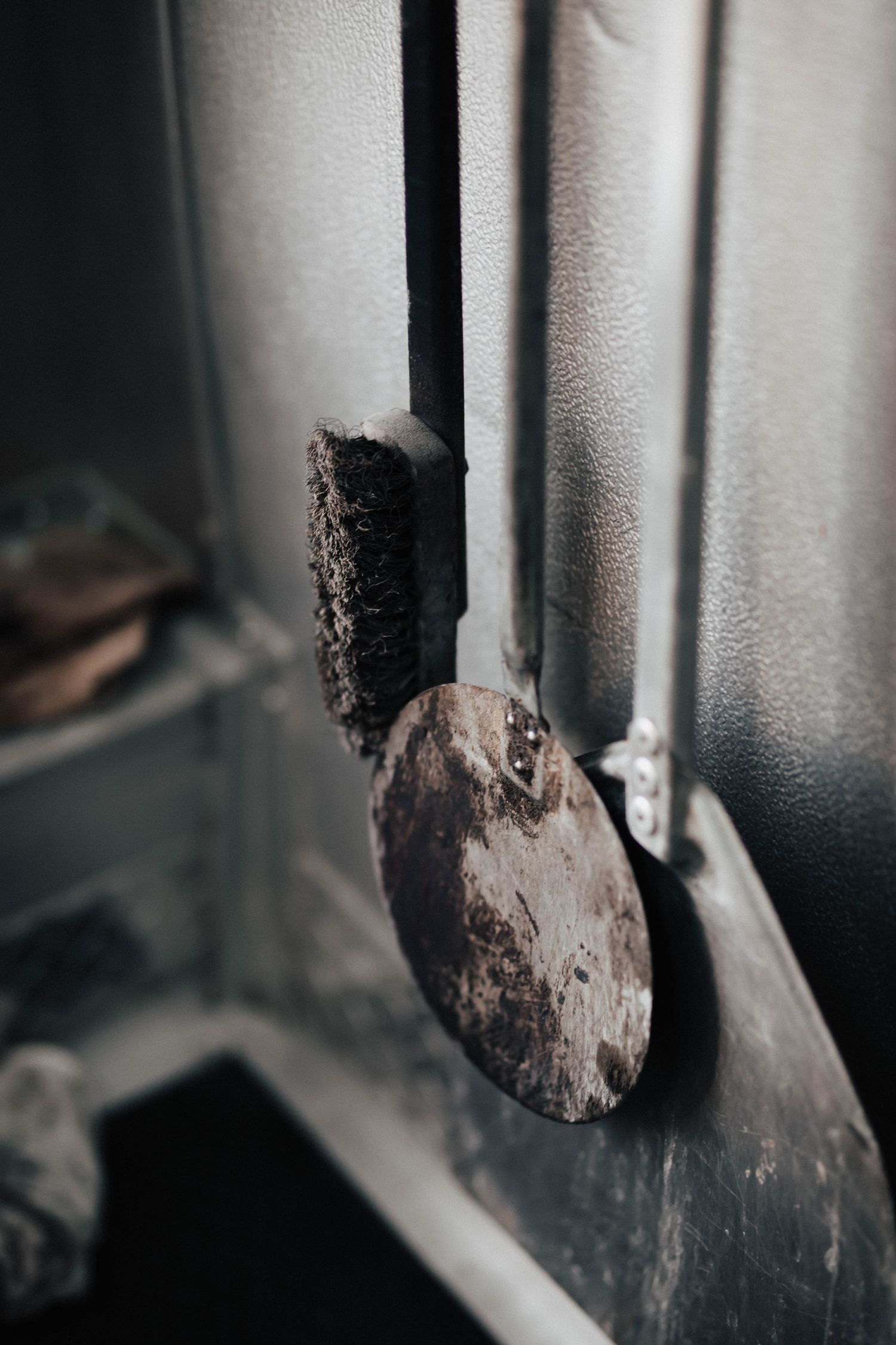 Worn wooden pizza peel brush hanging on the side of a black metal pizza oven.