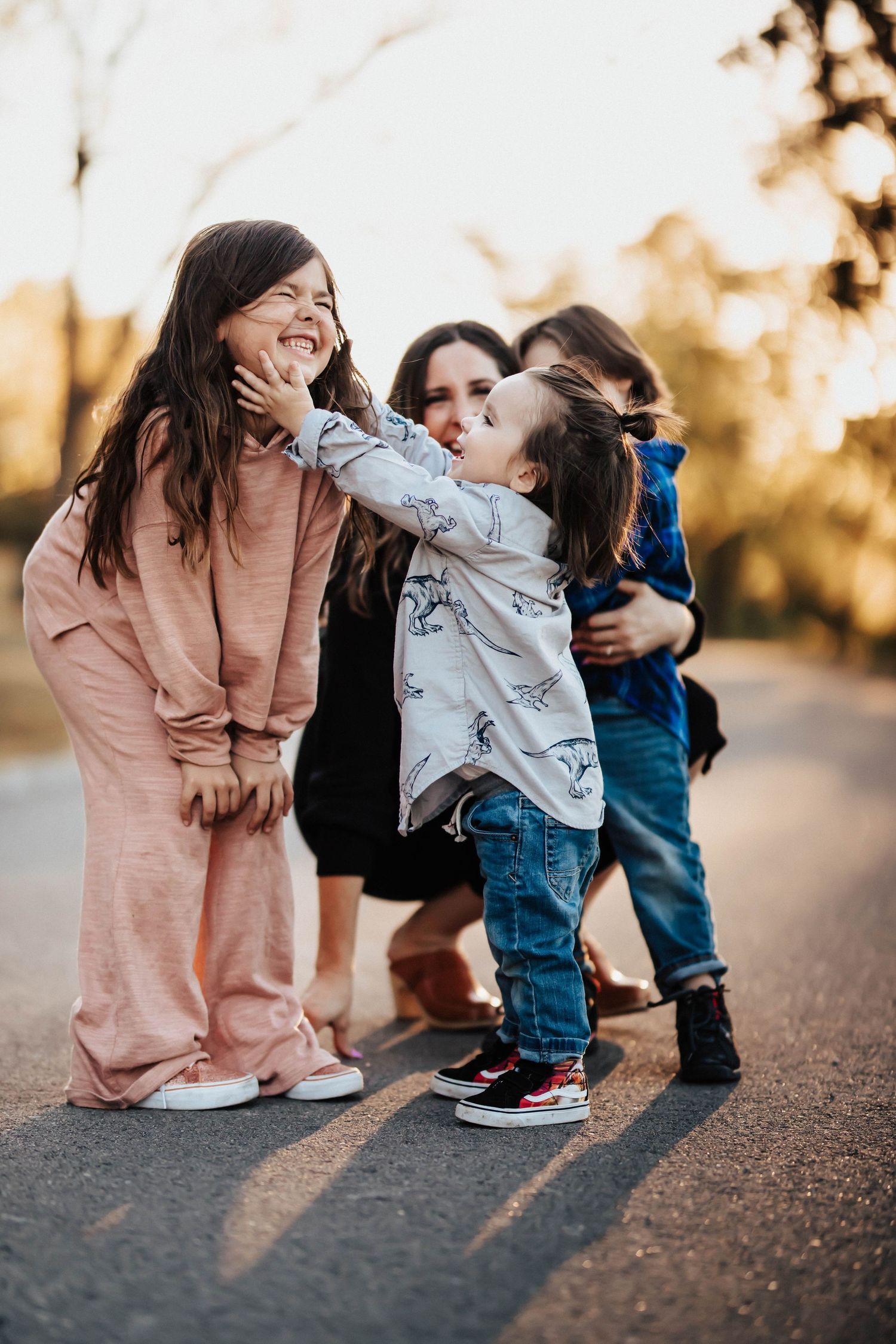 A mother crouching with three young children hugging and playing on a sunlit road.
