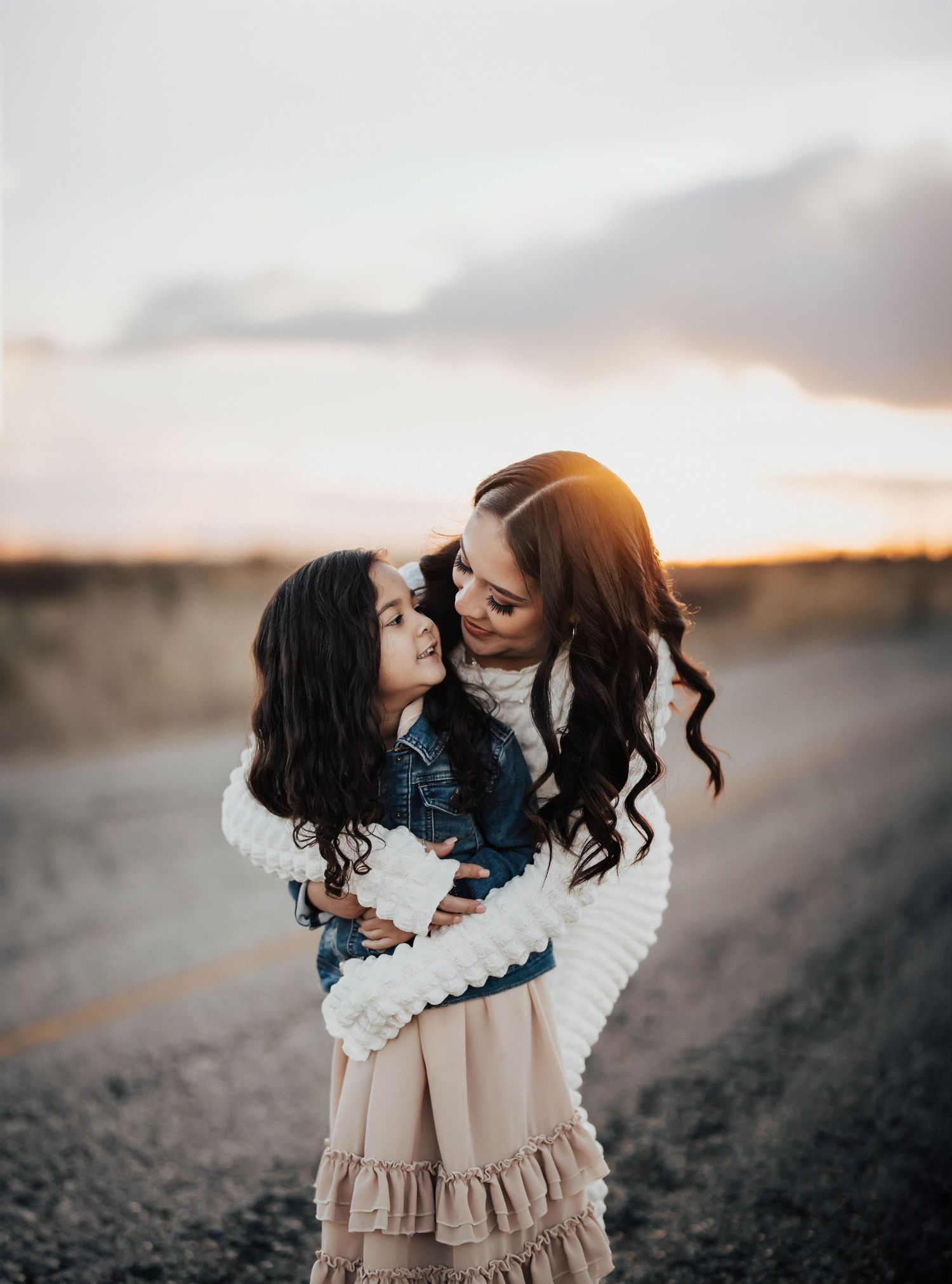 A mother tenderly embraces her young curly-haired daughter at sunset on an open road.