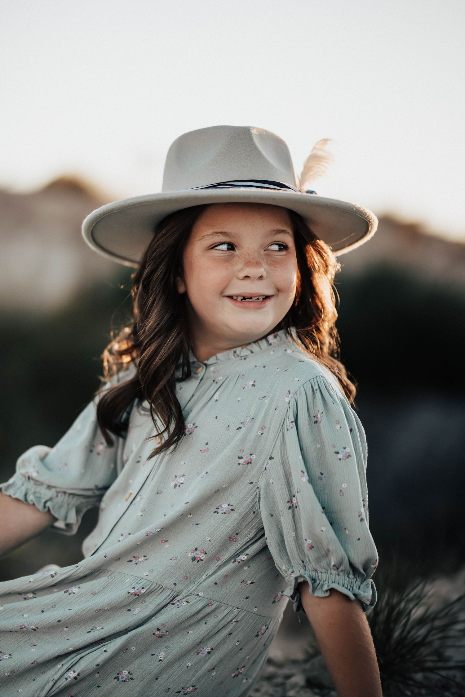 Cheerful young girl in floral dress and wide-brim hat smiling at sunset outdoor portrait.