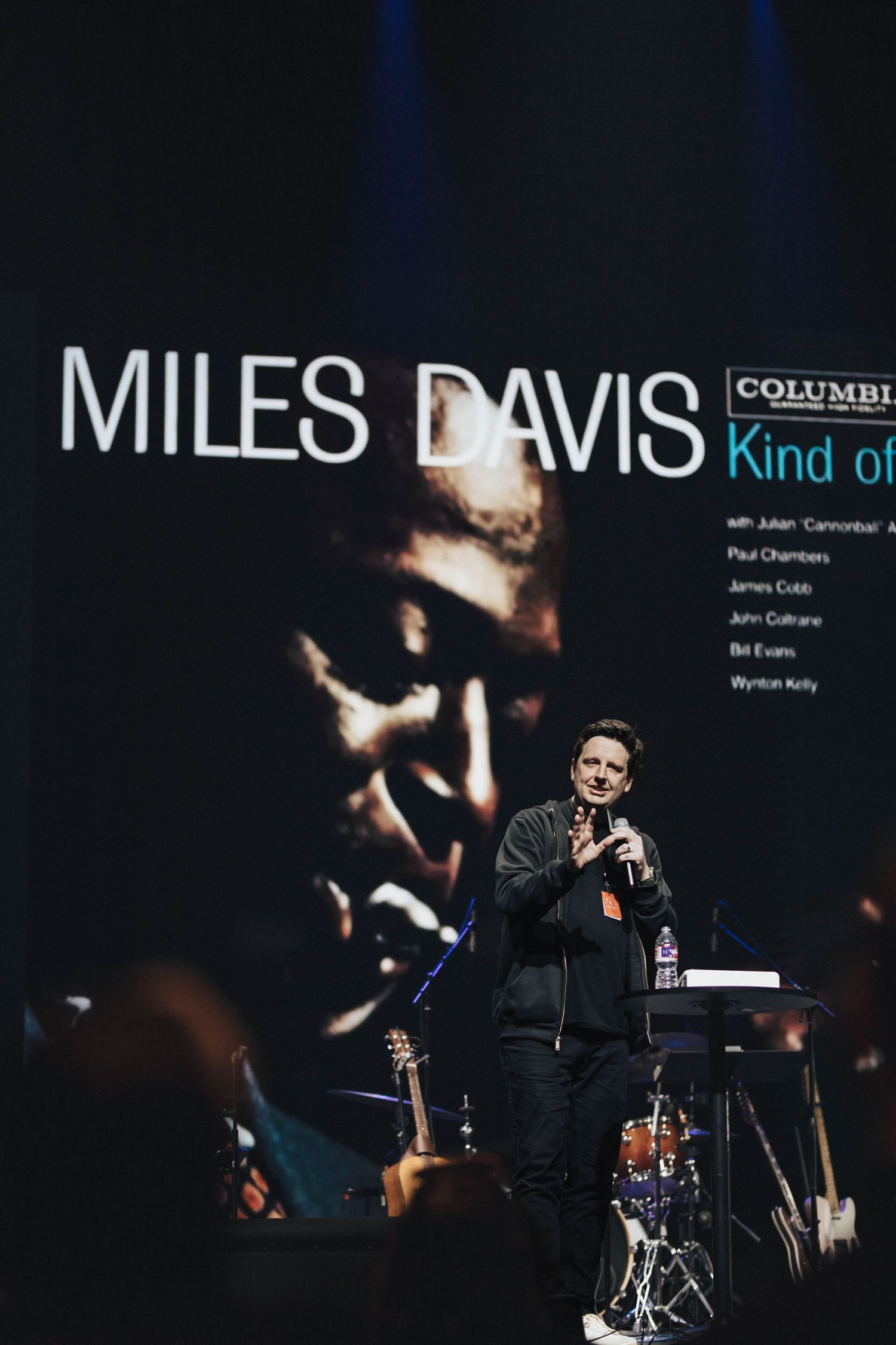 A speaker presents on stage in front of a Miles Davis Kind of Blue album cover projection.