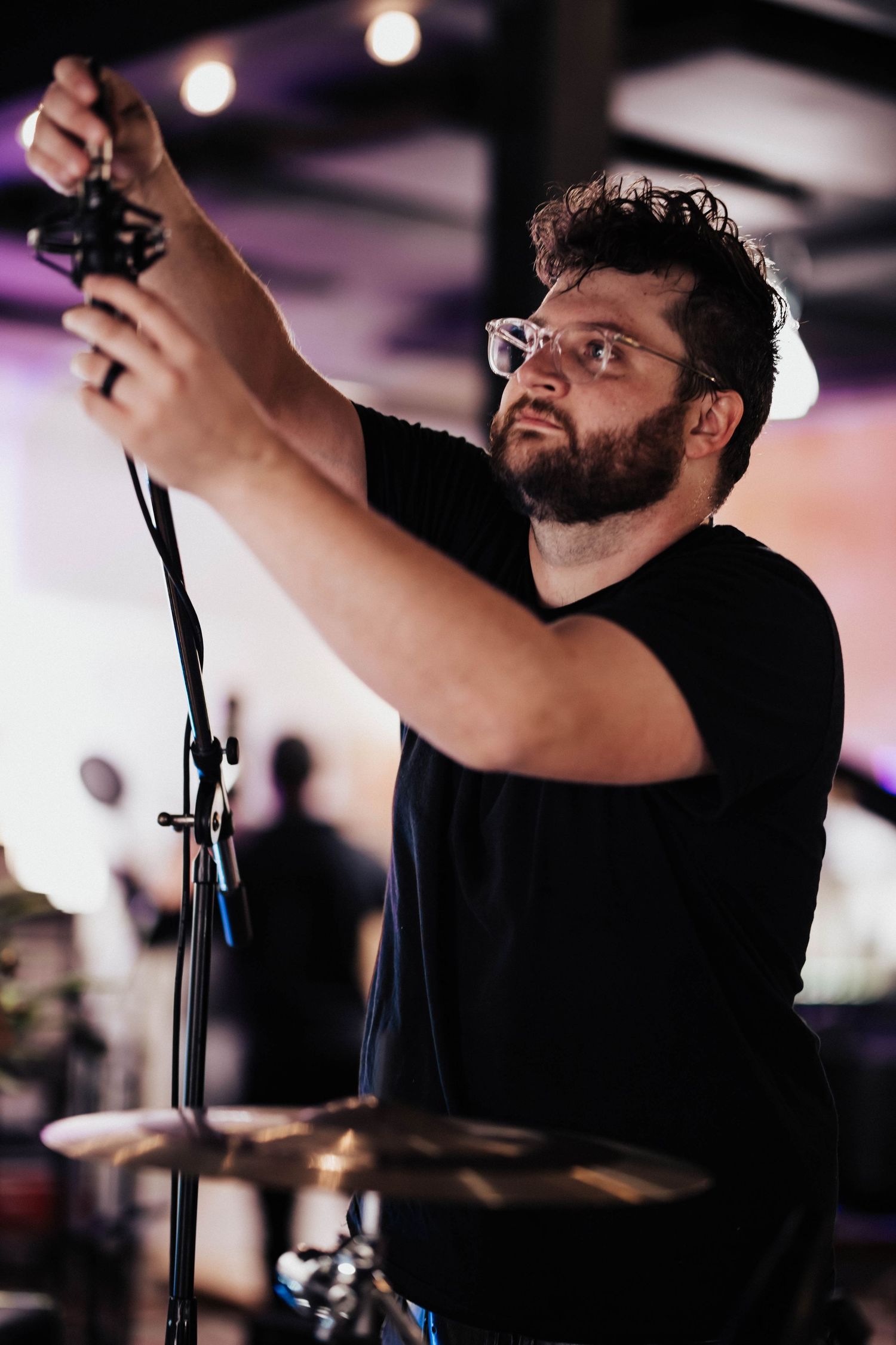 Bearded man with glasses adjusting a microphone stand near a drum kit at a live music event.