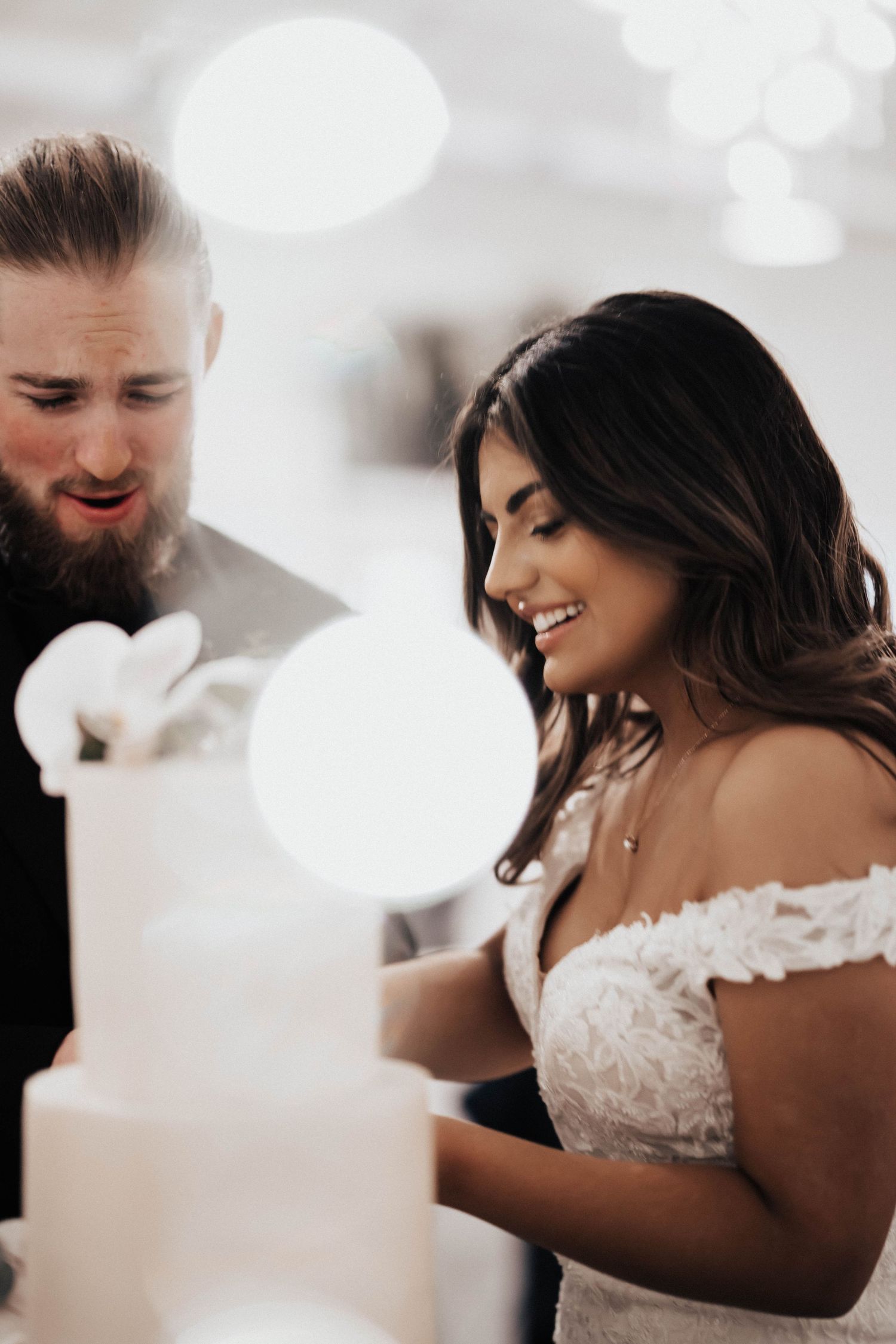 Bride and groom smiling while cutting their elegant white wedding cake during their reception ceremony.