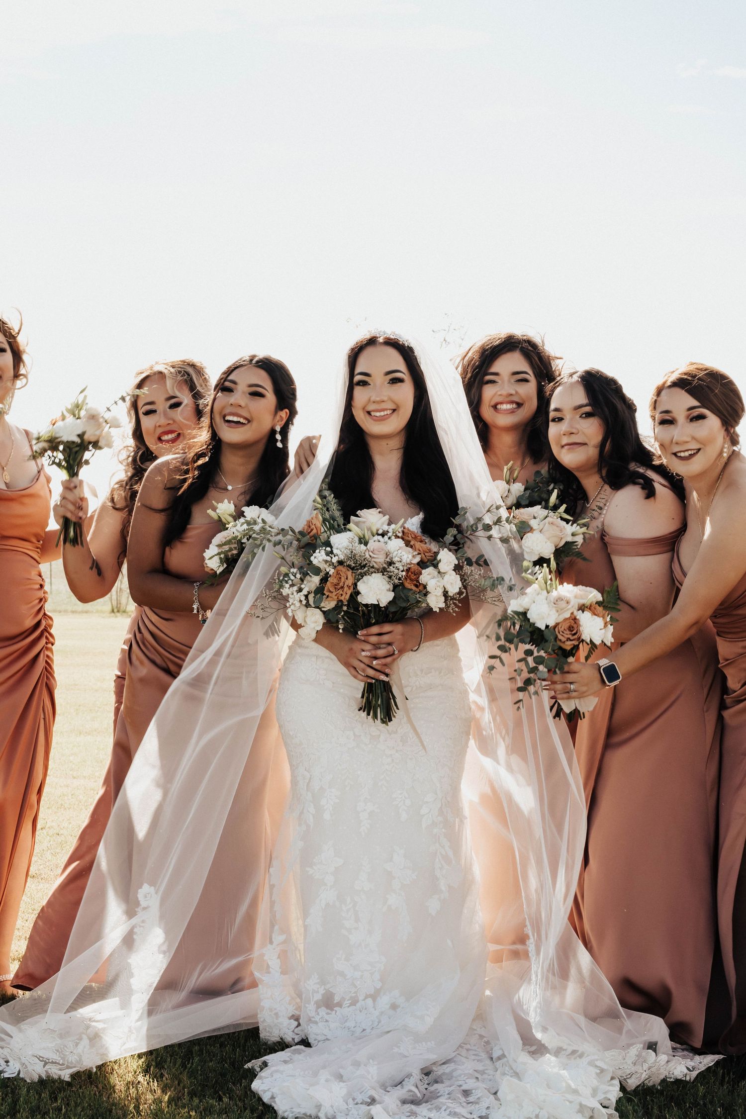 Bride in white lace gown with veil poses with six bridesmaids in dusty rose dresses holding bouquets outdoors.