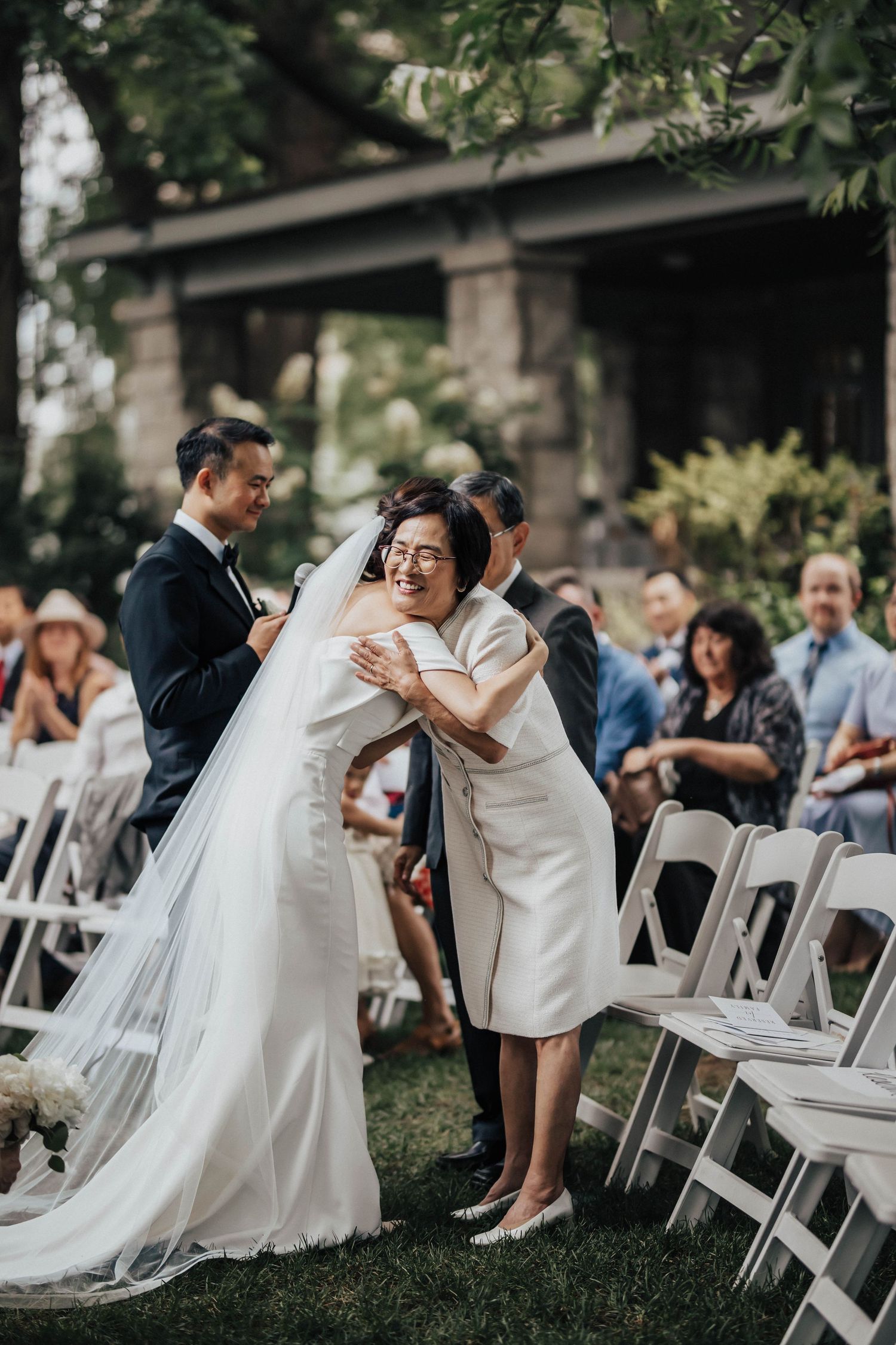 Bride in white gown hugs older woman in cream dress at outdoor wedding ceremony with white chairs and guests.