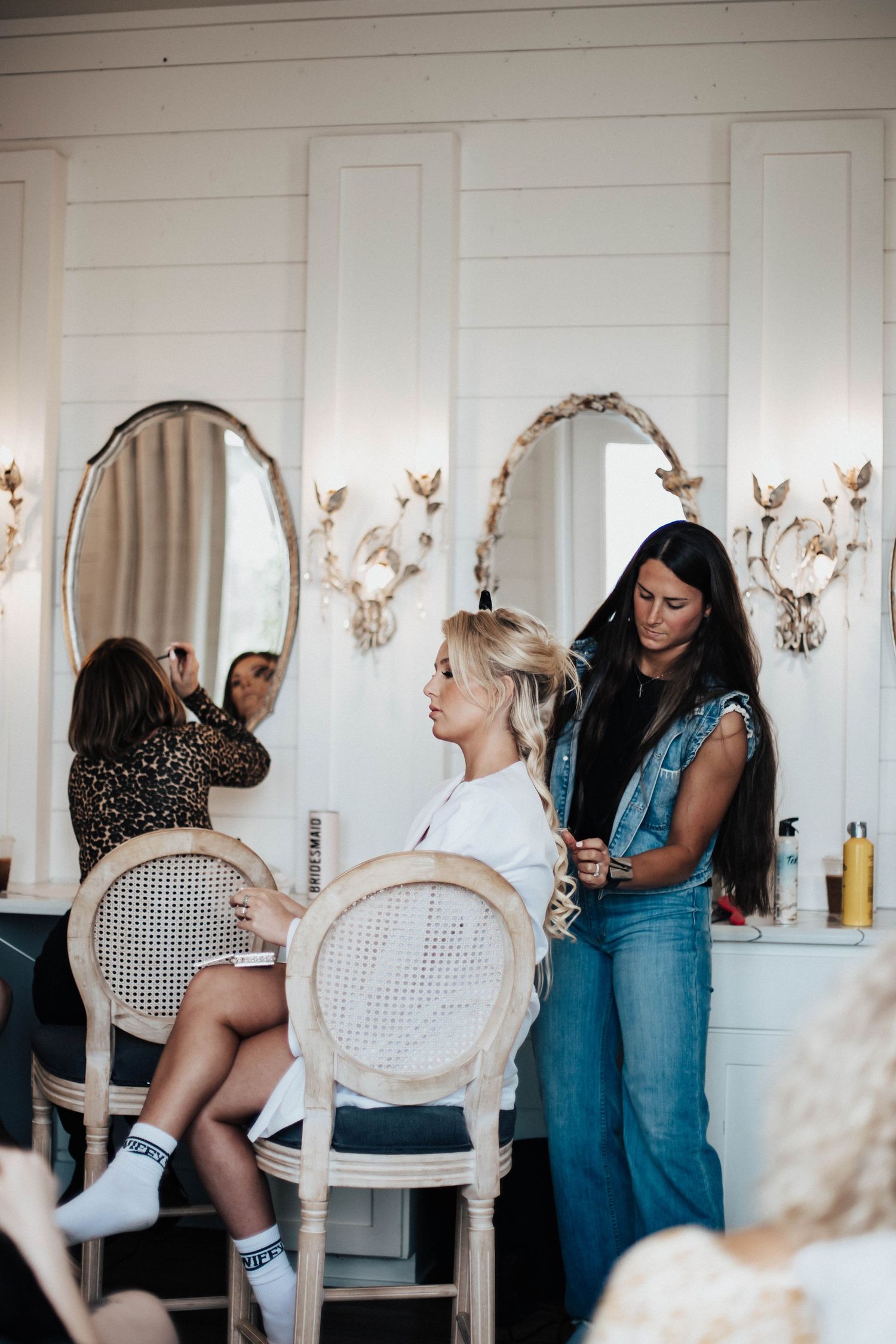 Bride gets hair styled by beautician in elegant white dressing room with oval mirrors before wedding.