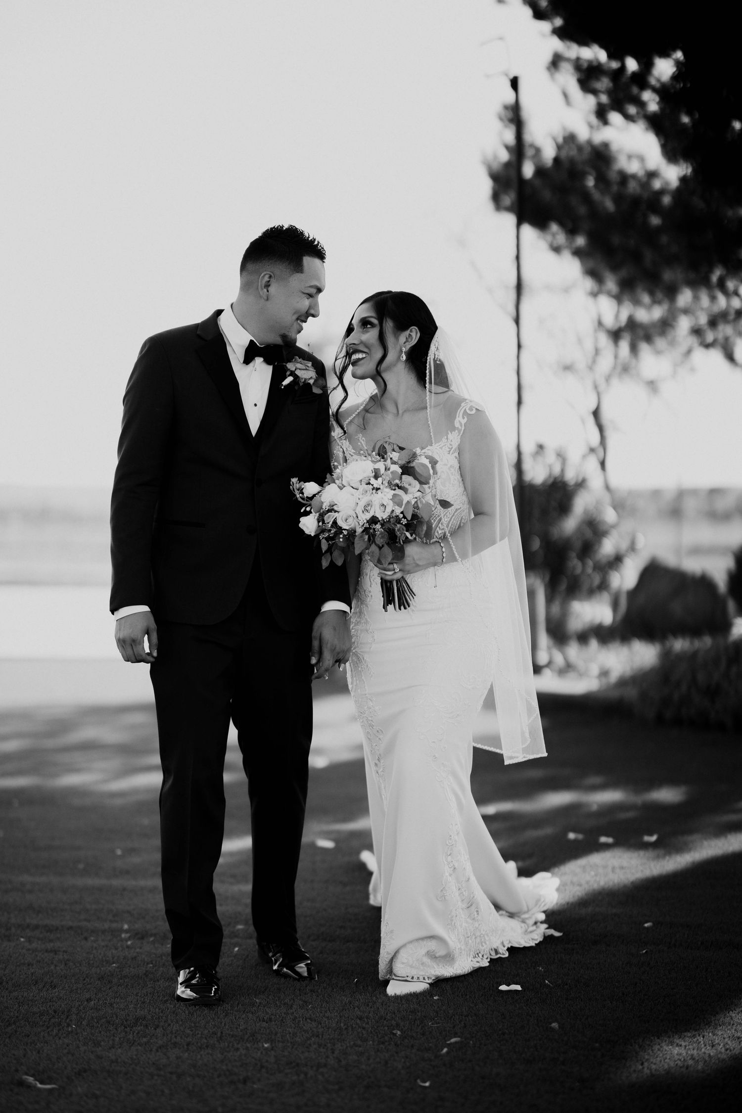 Black and white photo of a smiling bride and groom holding hands outdoors on their wedding day.