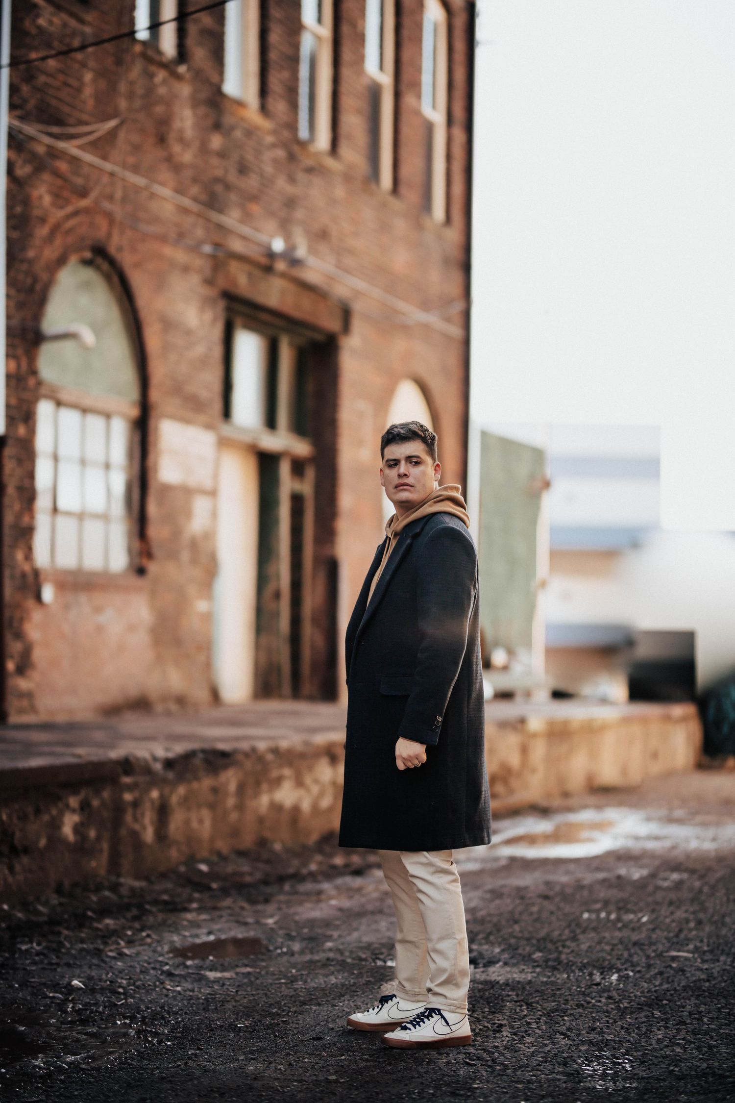Young man in black coat and tan hoodie stands on gravel lot beside brick building.