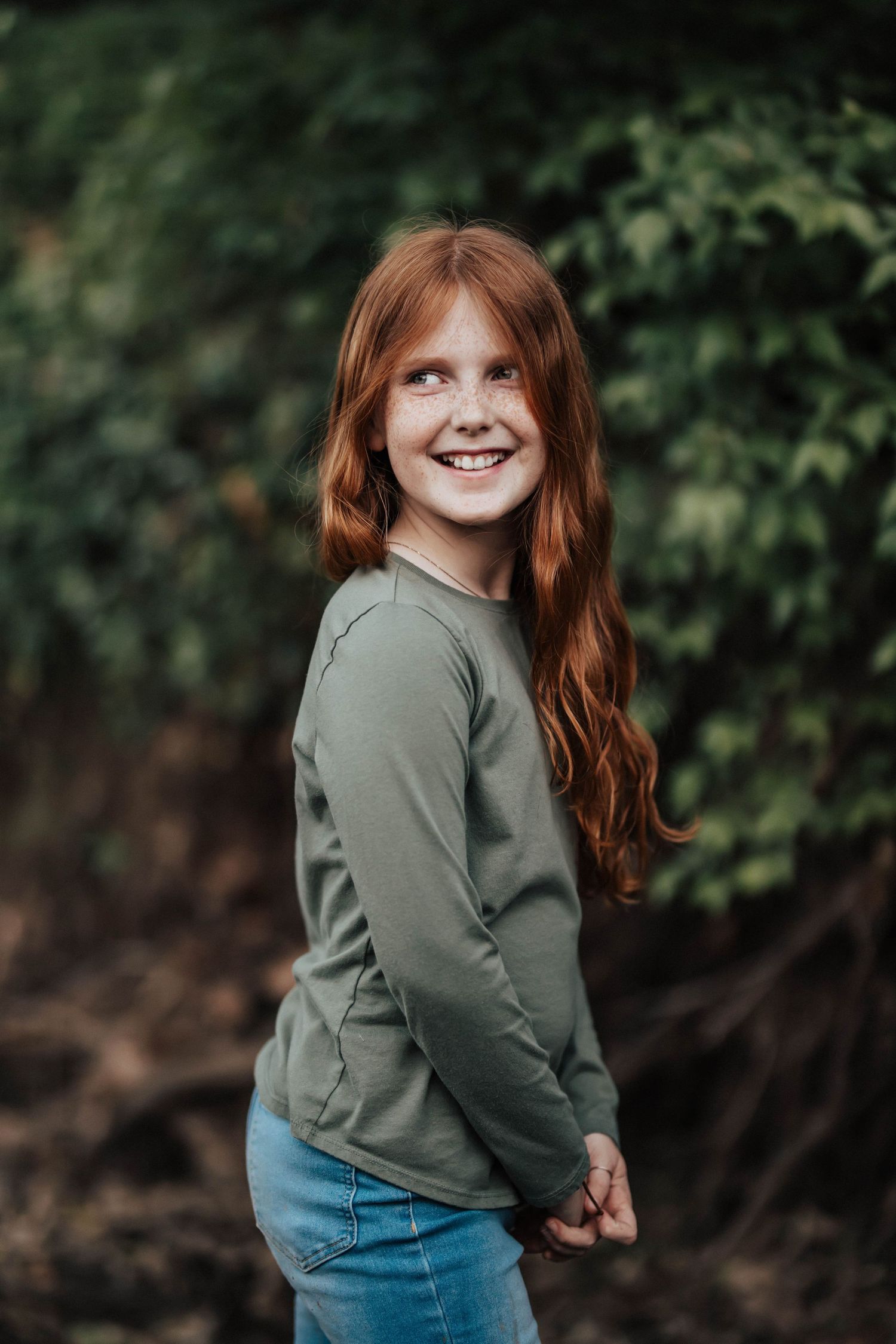 Smiling redhead girl with freckles in green sweater and jeans posing outdoors among lush green foliage.