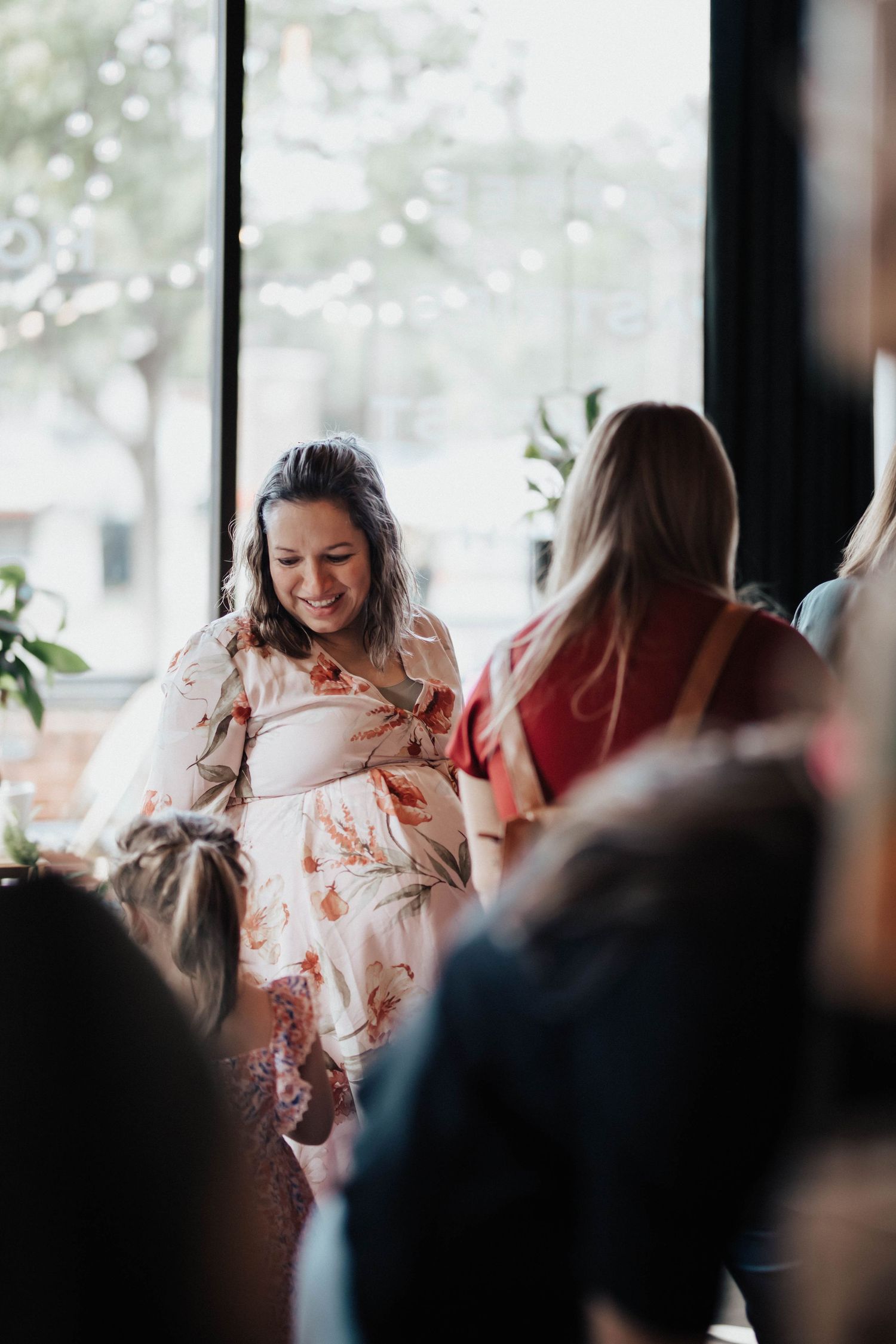 Smiling pregnant woman in floral dress stands near large windows at a baby shower.