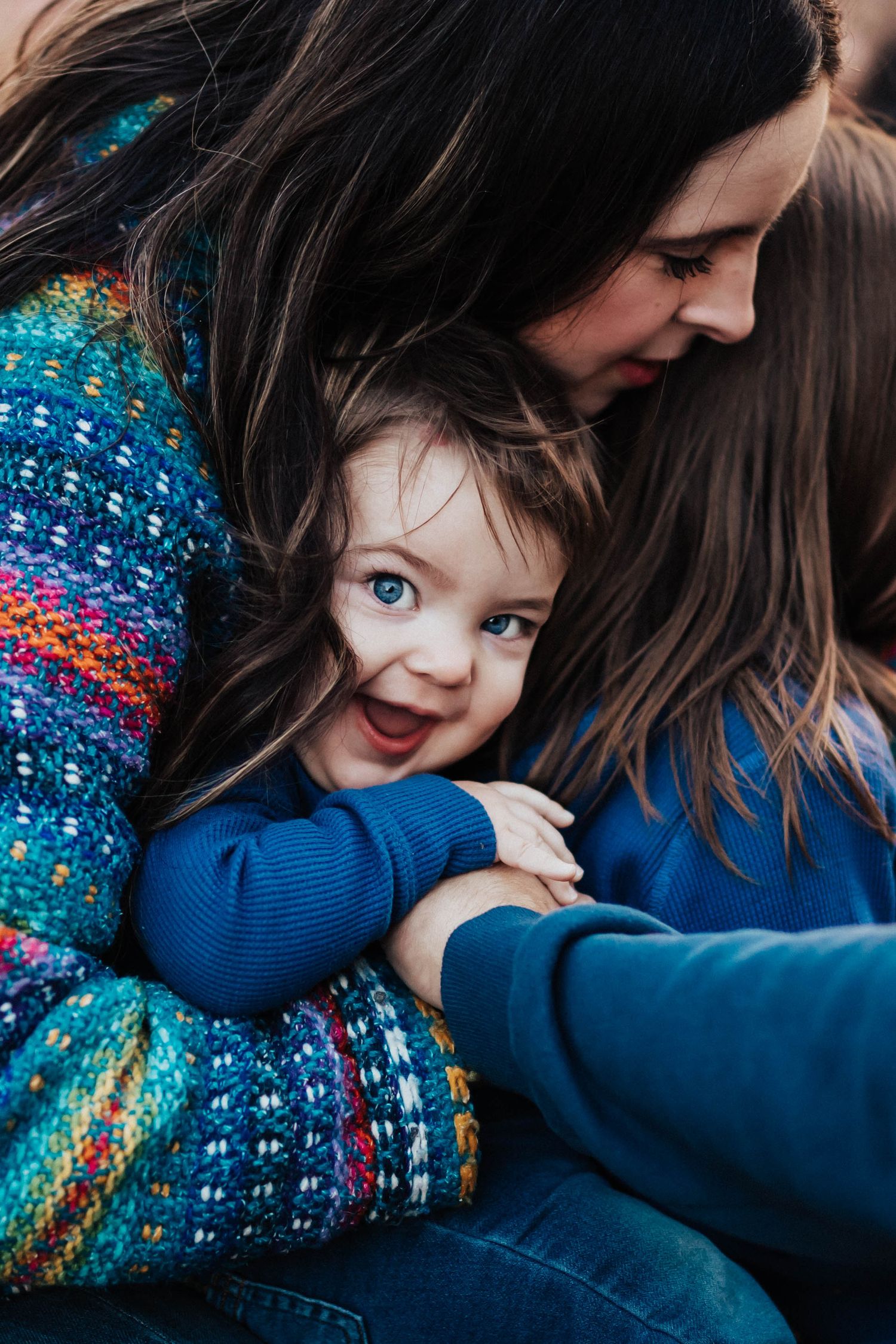 A woman in a colorful sweater hugs a bright-eyed smiling toddler with blue eyes warmly.