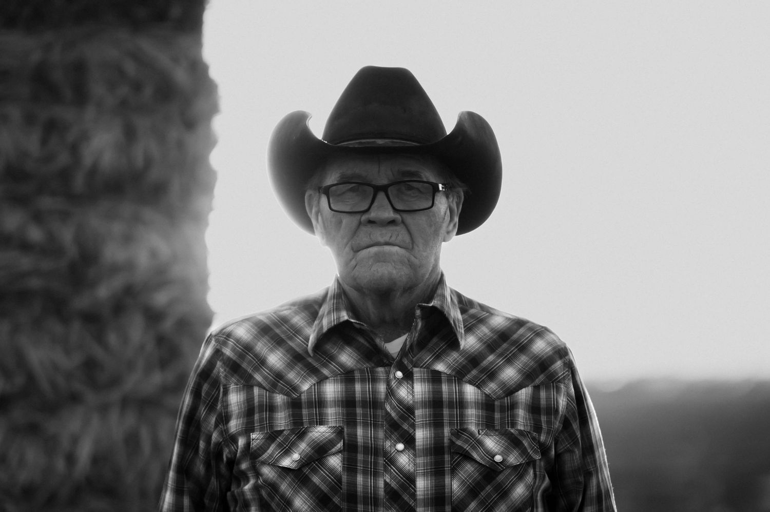 Black and white portrait of an elderly man wearing a cowboy hat and plaid shirt standing outdoors.