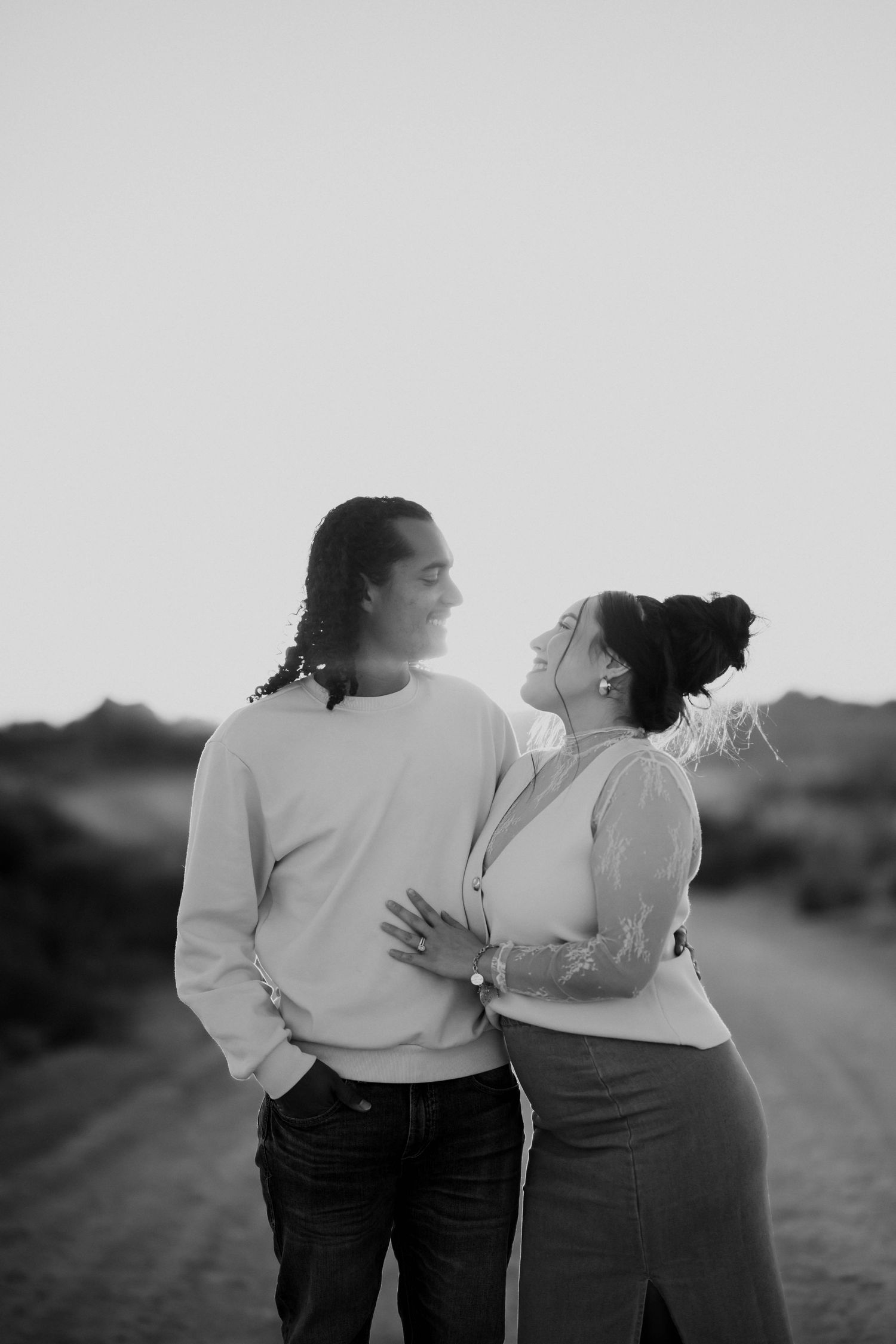 Black and white photo of couple standing on dirt road at sunset, woman embracing man from behind.