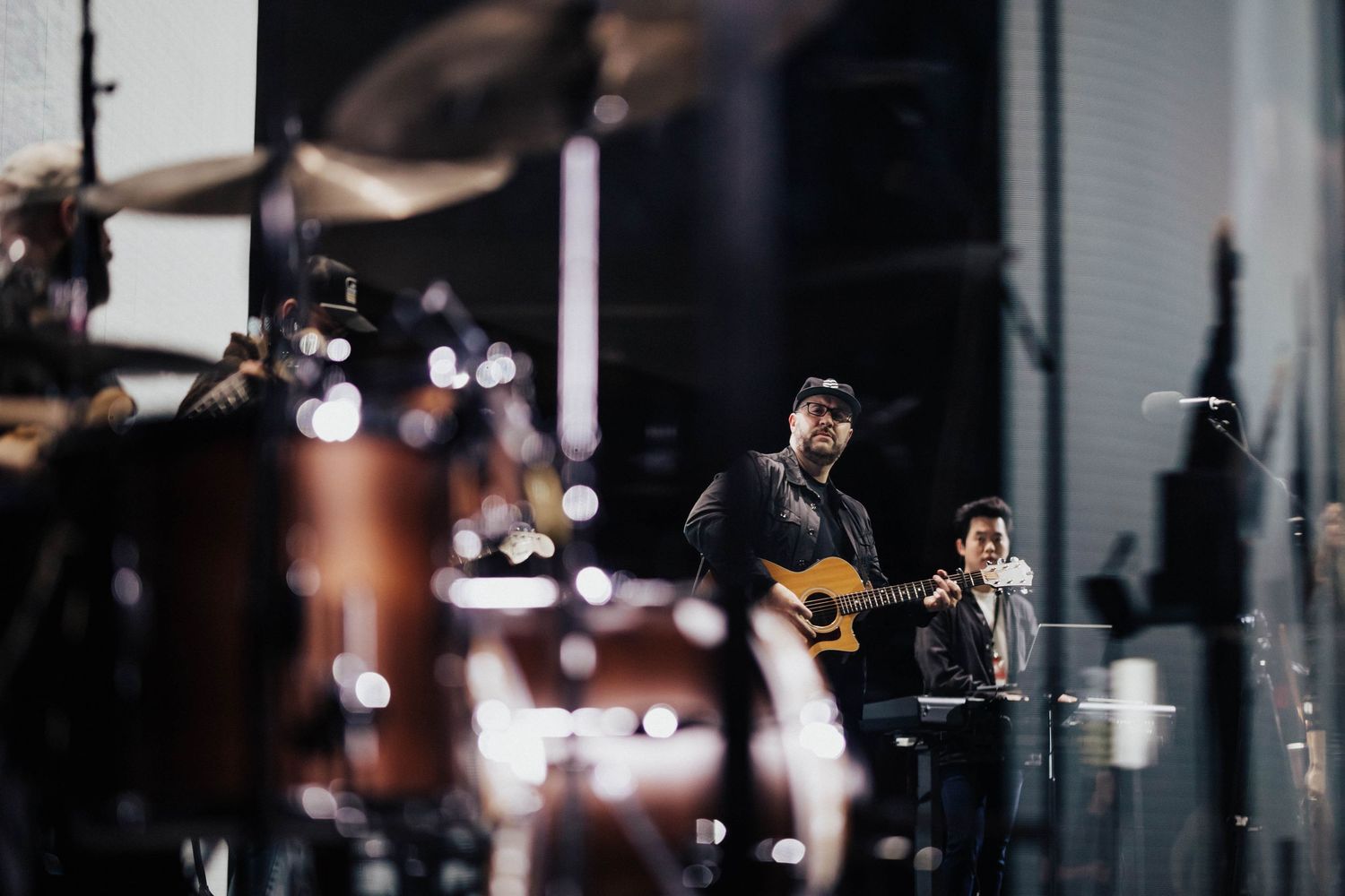 Two musicians perform on stage, one playing acoustic guitar, with a drum kit visible in the background.