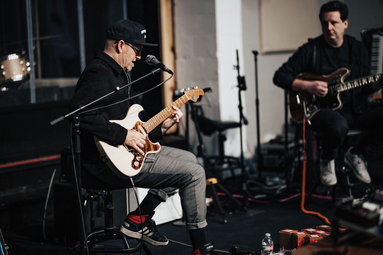 Musician in black cap plays electric guitar seated on stage during rehearsal with second guitarist behind him.
