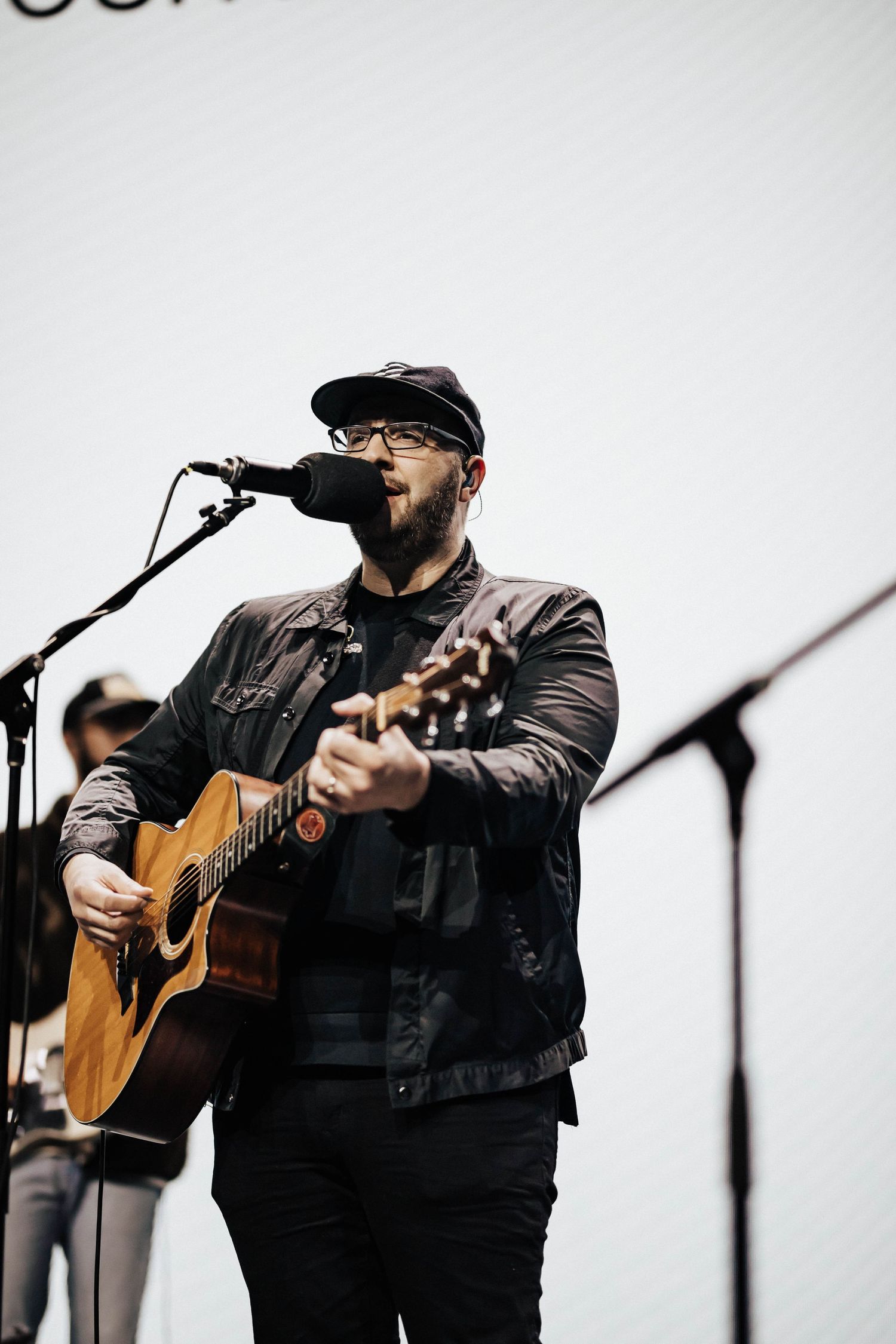 Bearded male musician playing acoustic guitar and singing into microphone on stage wearing black jacket and cap.