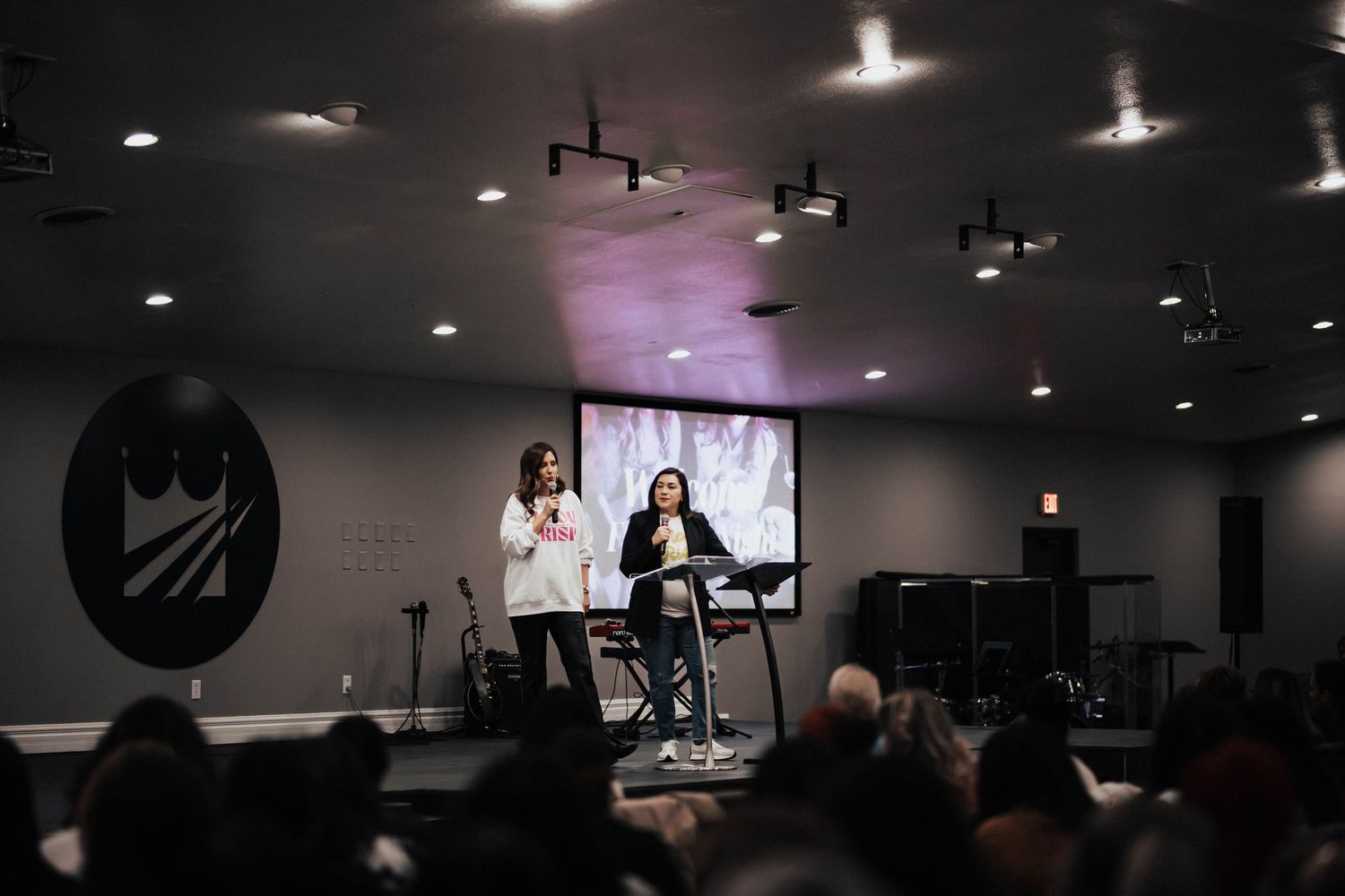 Two women speak on stage at a church event with a screen projection behind them.