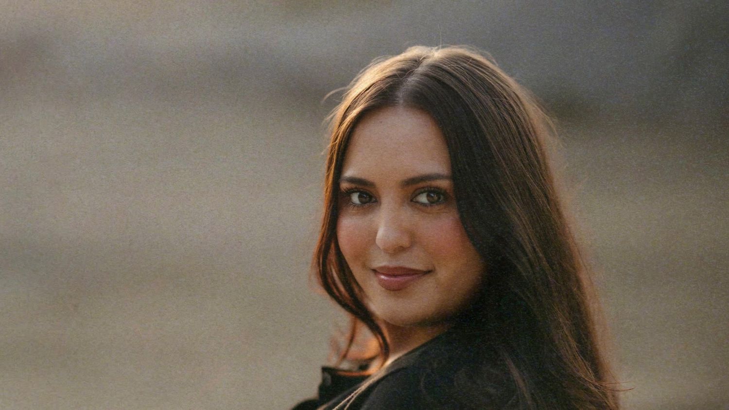 Young woman with long dark hair smiling over her shoulder in a golden hour outdoor portrait.