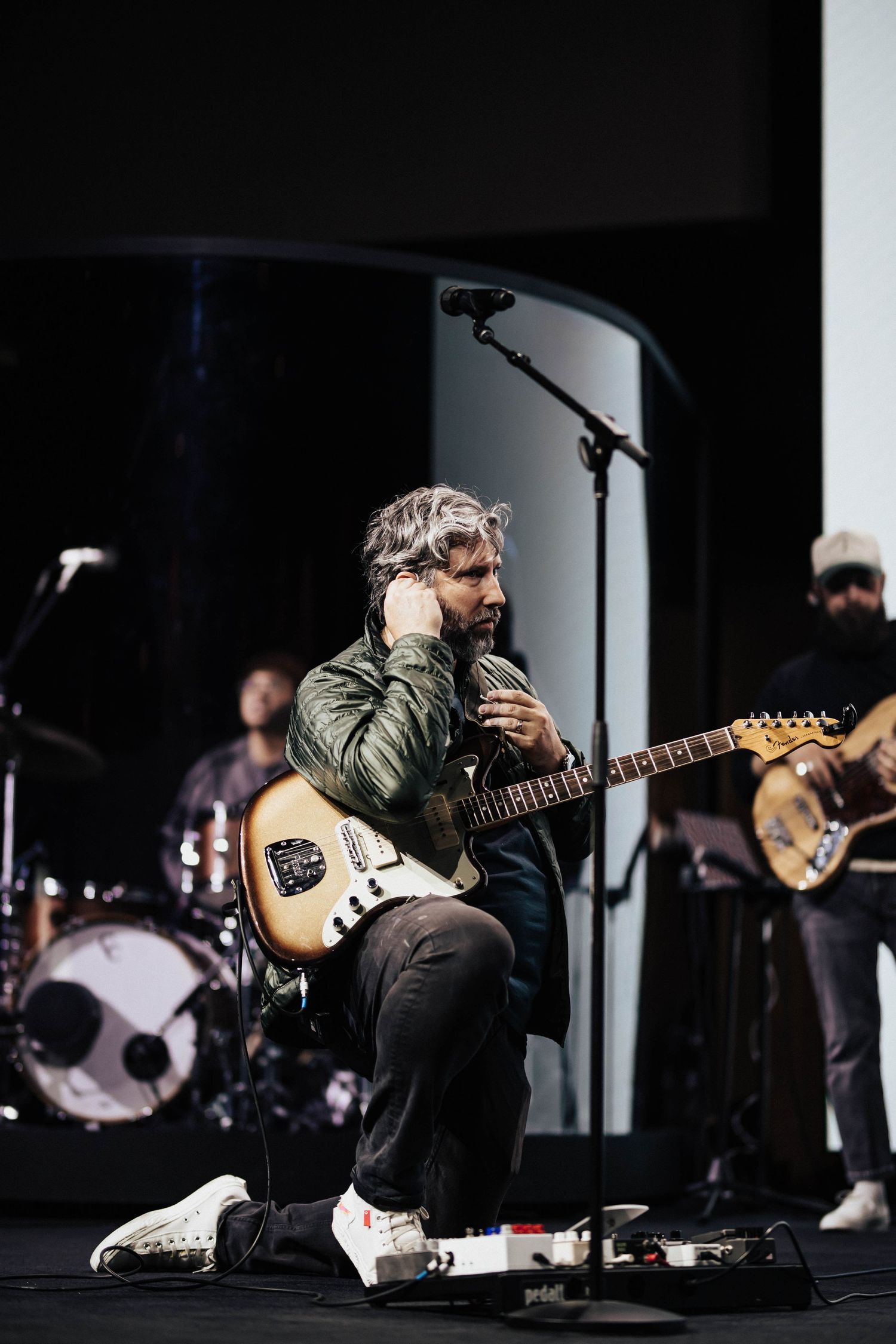 Gray-haired guitarist kneeling on stage with band members and drum kit visible in background.