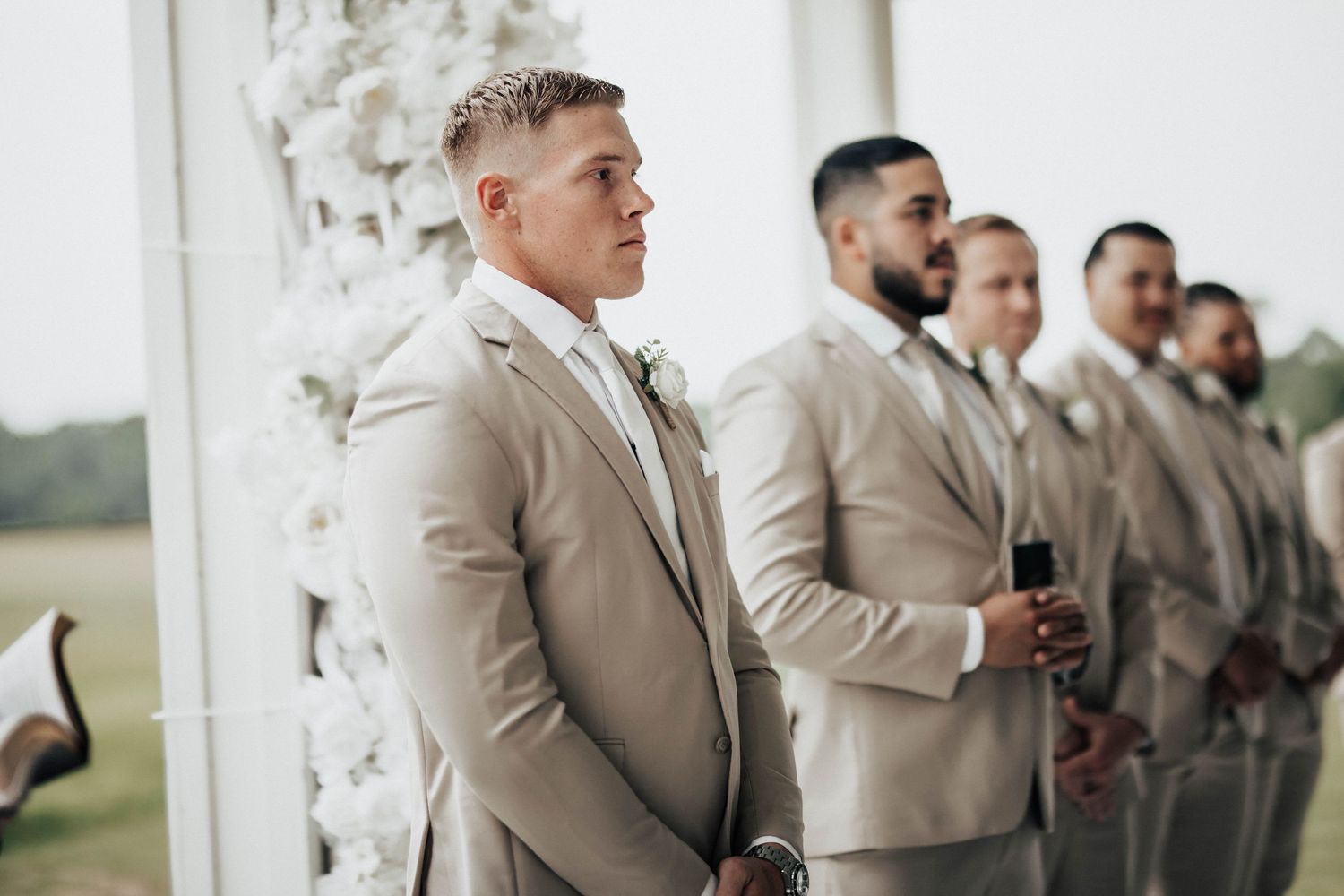 Groom in tan suit stands with groomsmen at wedding altar decorated with white floral arrangements.