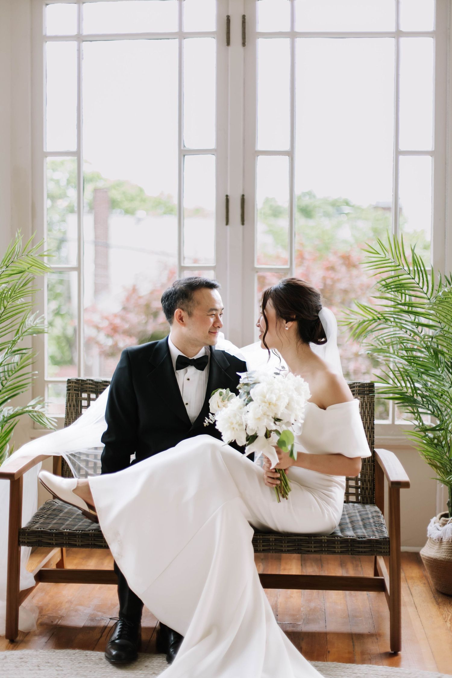 Bride and groom sit together on a bench, exchanging loving glances, bride holding white bouquet in elegant wedding attire.