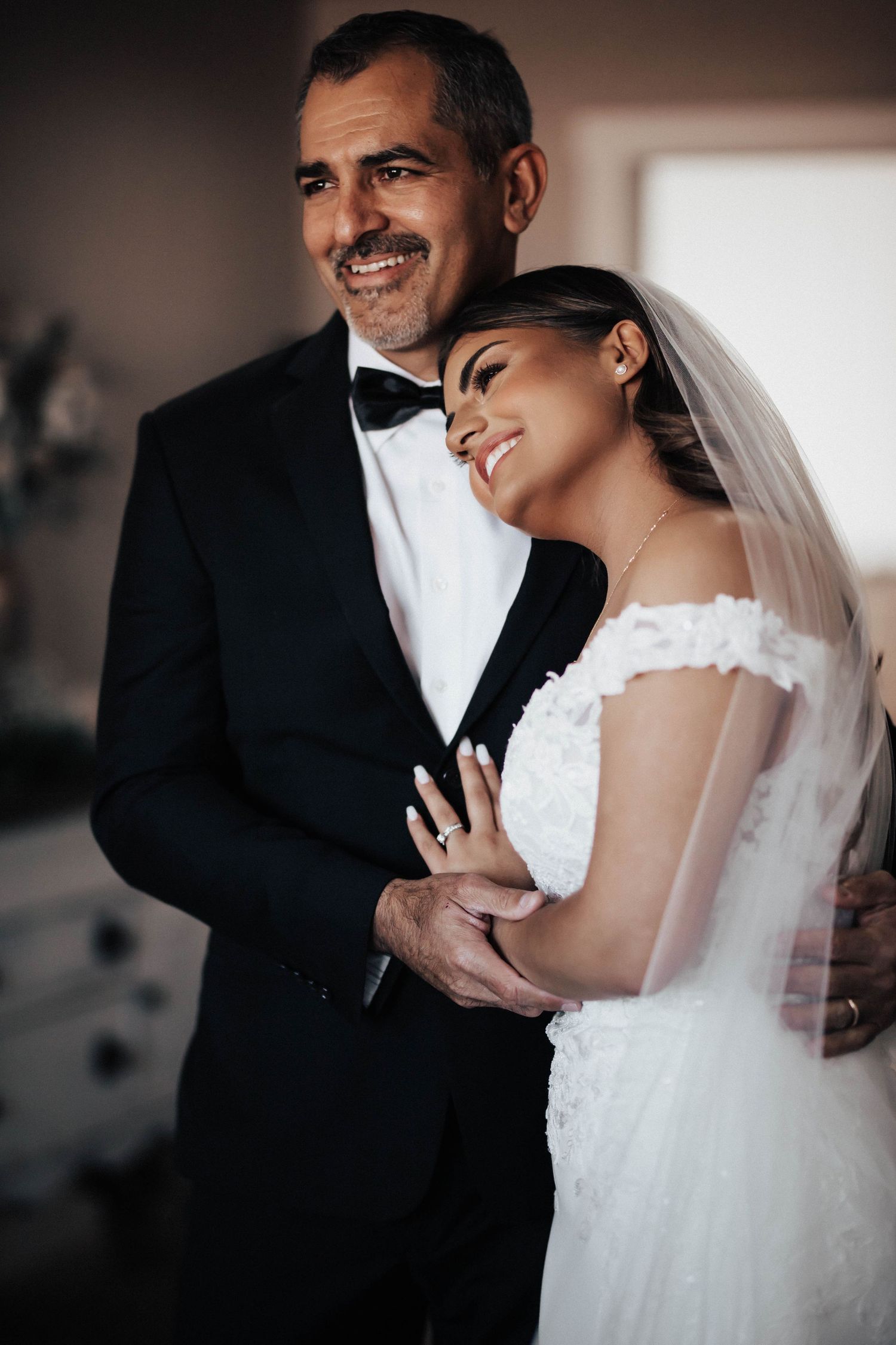 A smiling bride in a white lace off-shoulder gown and veil embraces a groom in a black tuxedo with bow tie.