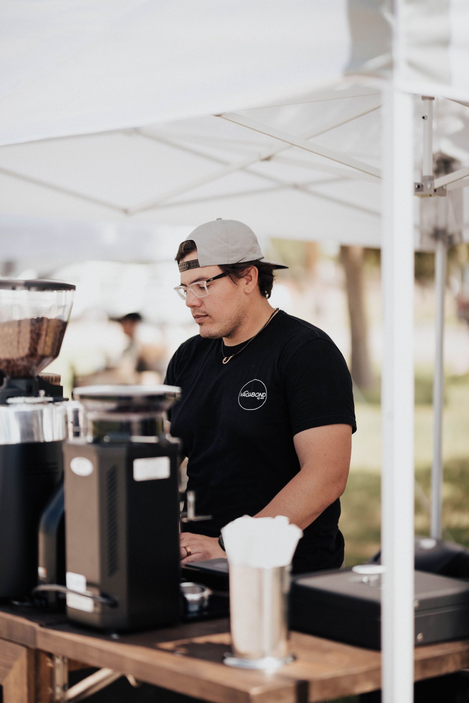 Barista in black Vagabond Coffee shirt operates commercial grinders at outdoor coffee stand.