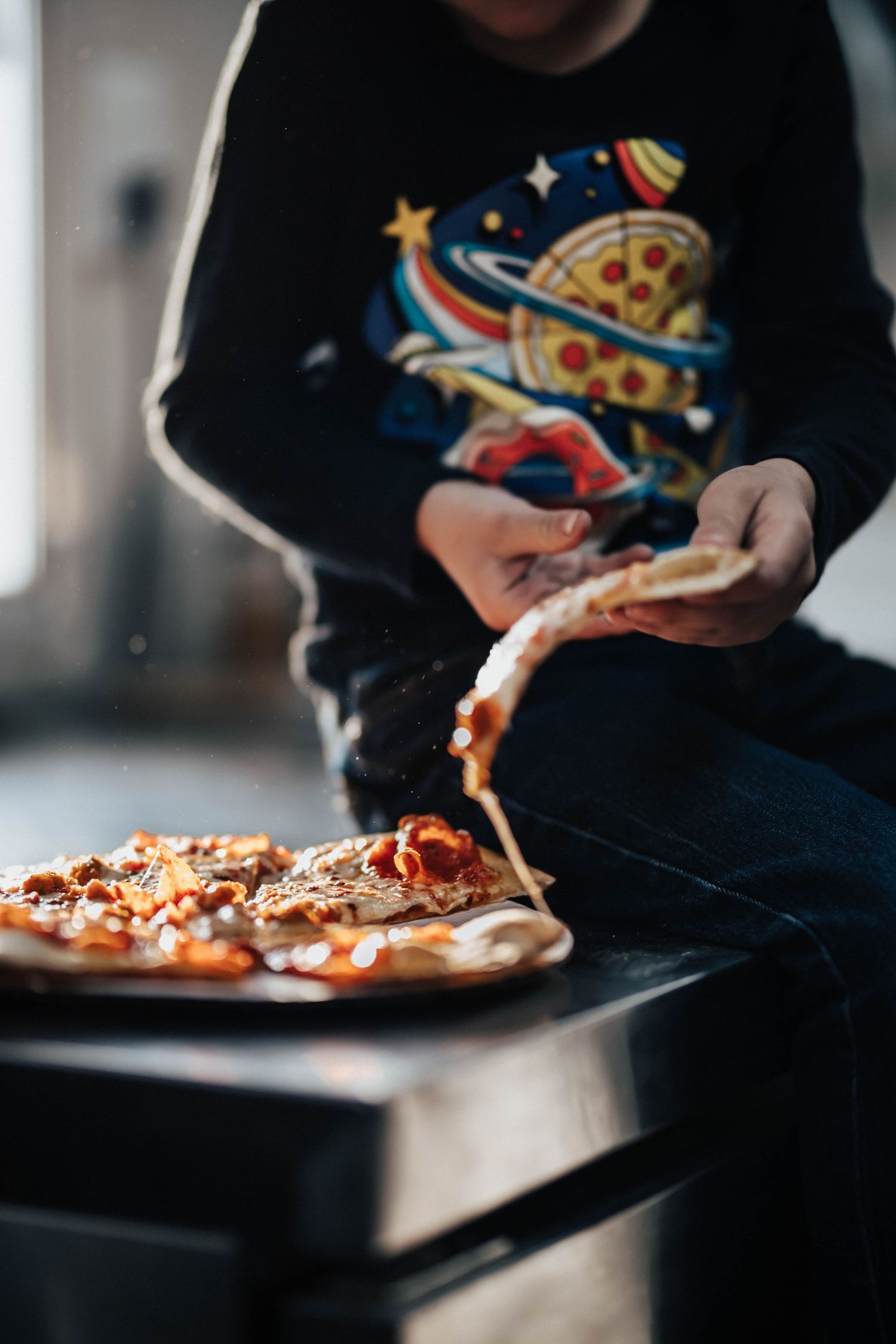 Child in a space-themed sweatshirt pulling a cheesy slice from a whole pizza.