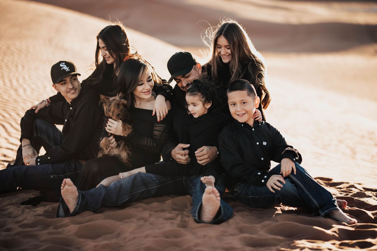 A happy family of seven sits together casually on sandy desert dunes with a small dog.