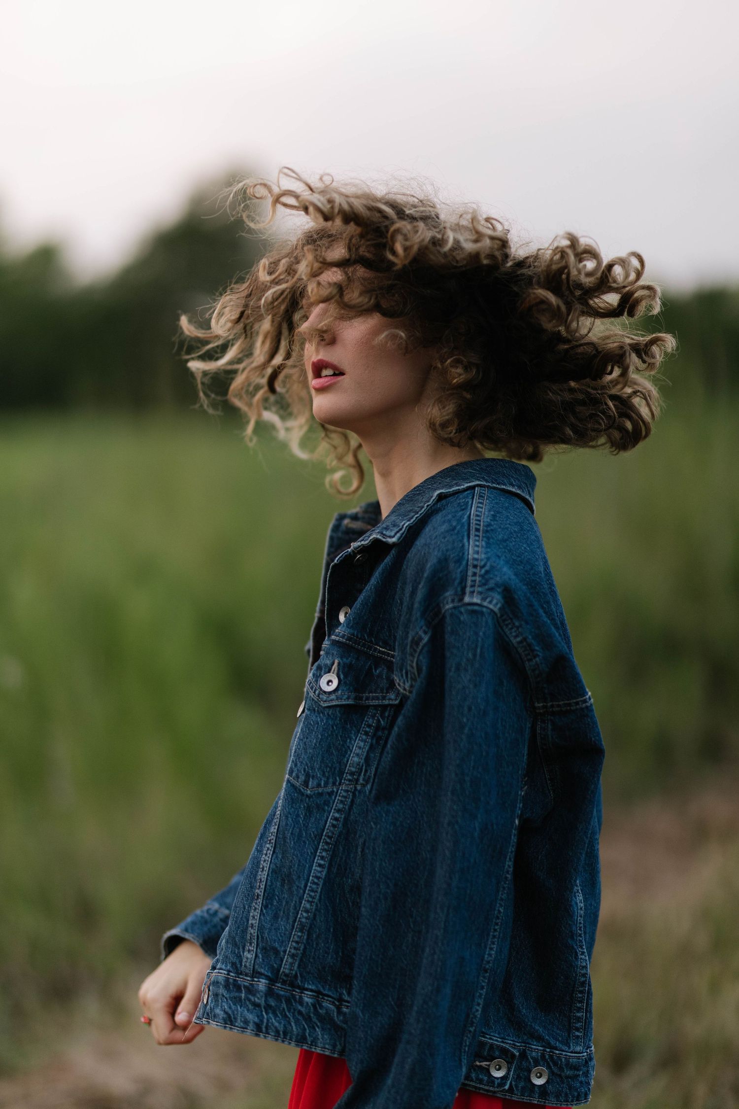 Young woman with curly hair tossing her head in a green field wearing a denim jacket and red pants.
