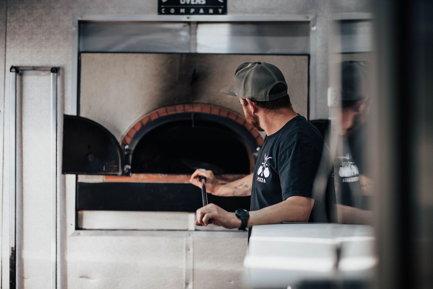 A pizza chef working at a wood-fired brick oven in a professional kitchen, wearing a black t-shirt and cap.