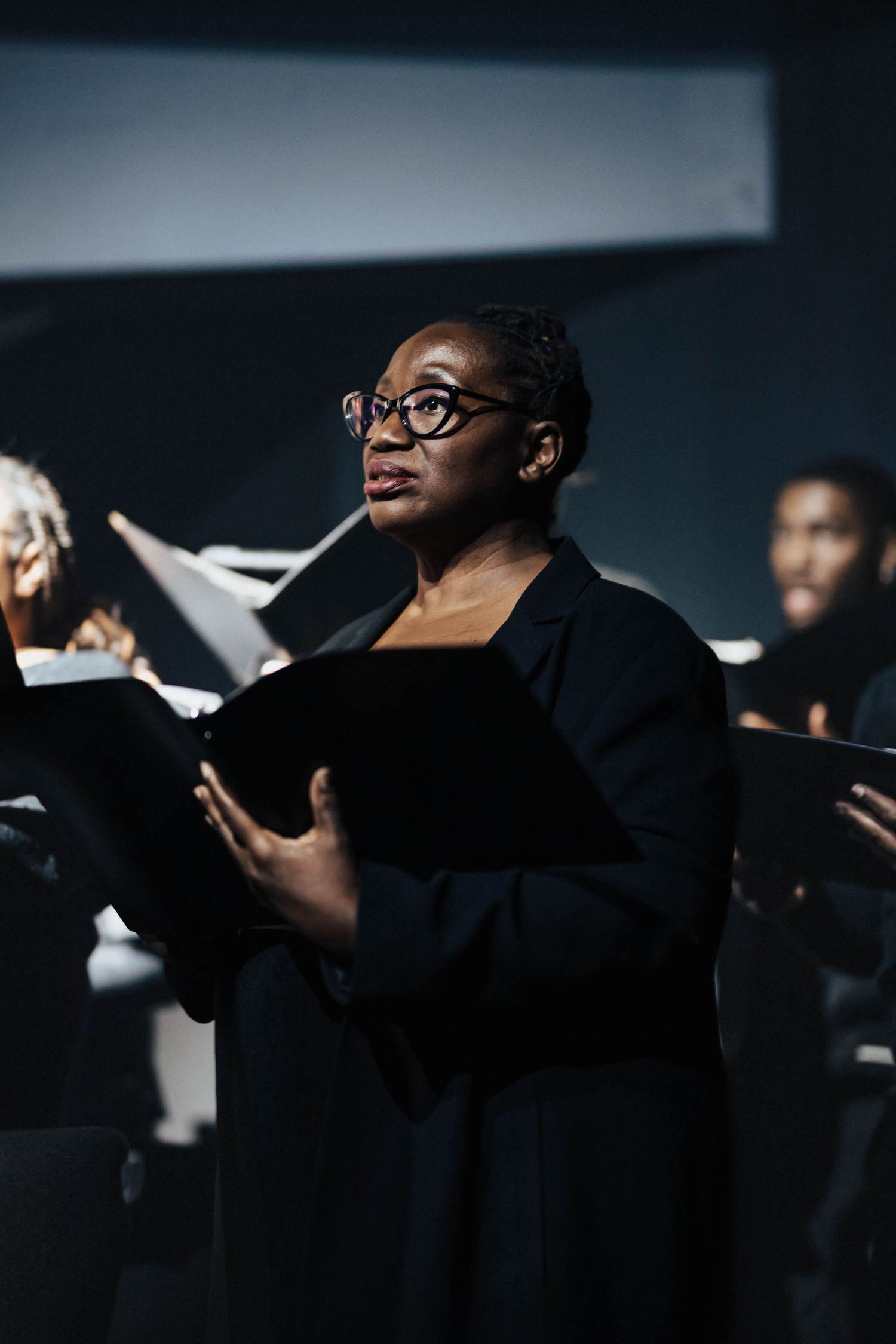 Black woman in glasses holding sheet music folder performing with choir in dark stage setting.