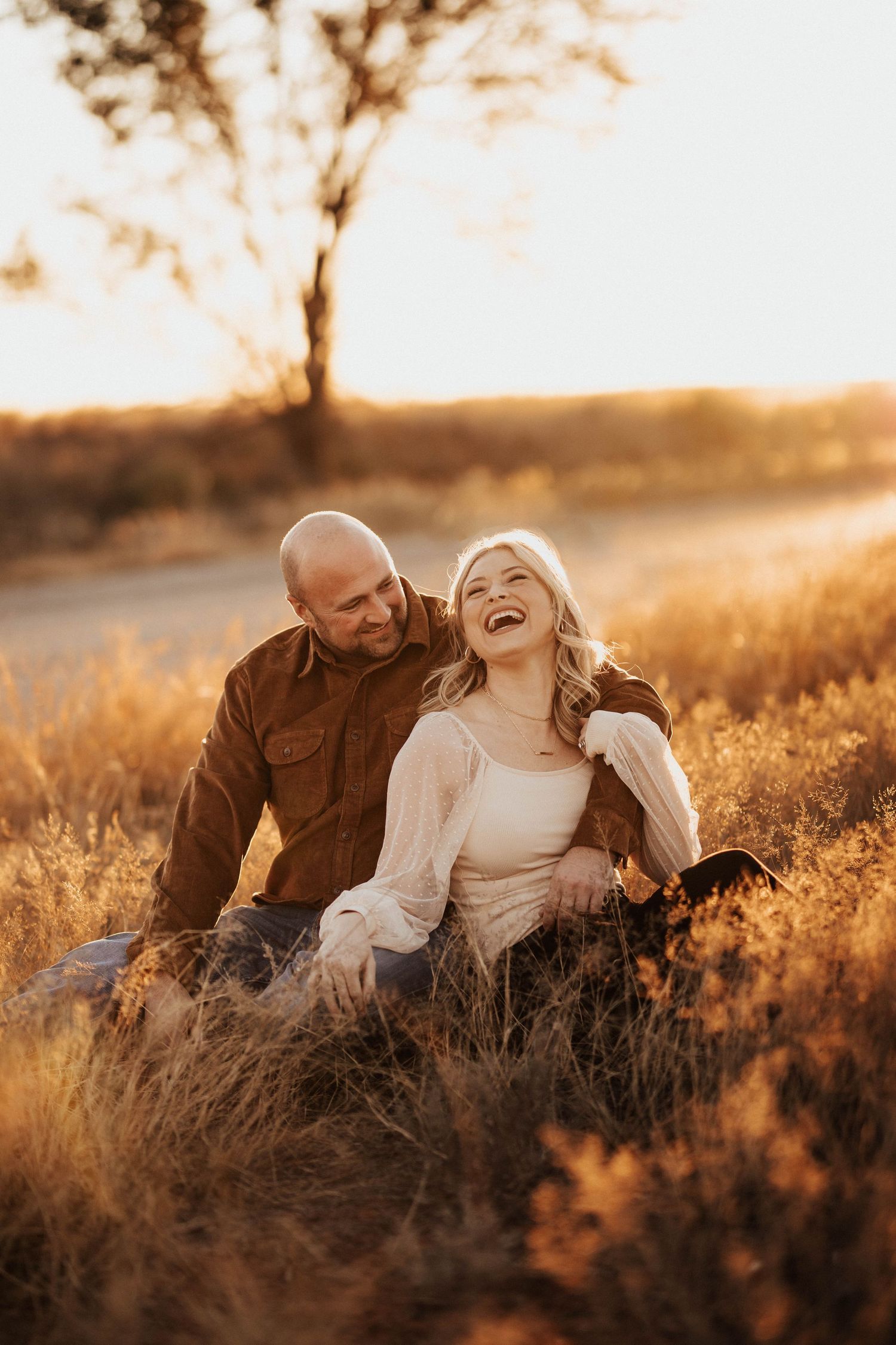 Laughing couple sitting together in golden autumn grass at sunset, woman in white sweater.